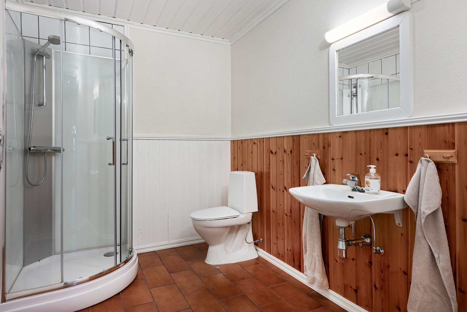 Badet i underetasjen er fra byggeår med flislagt gulv og panelte vegger, og er innredet med dusjkabinett, gulvmontert toalett, servant og speil. Galleribilde