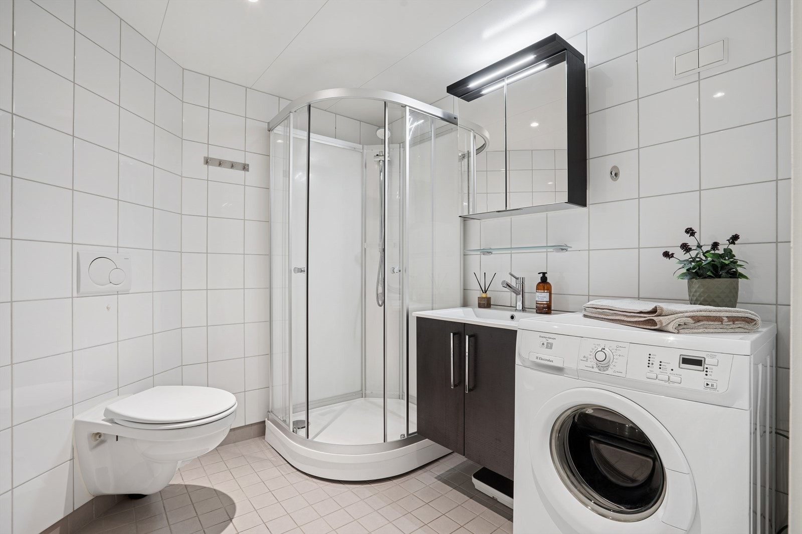 Flislagte vegger og elektriske varmekabler gir robust våtromsløsning. Badet har mekanisk avtrekk og inkluderer dusjkabinett, veggmontert toalett og opplegg for vaskemaskin. Galleribilde