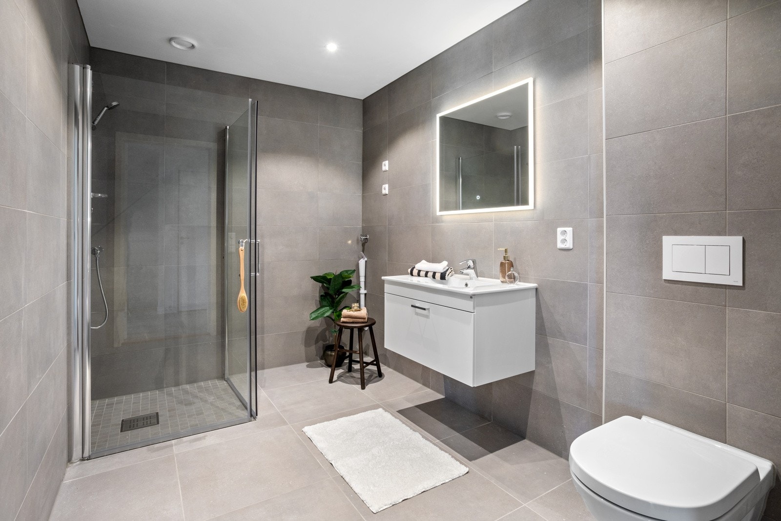 Moderne baderom med dusjnisje, vegghengt toalett og servant med speil. Flislagte vegger og gulv. Galleribilde