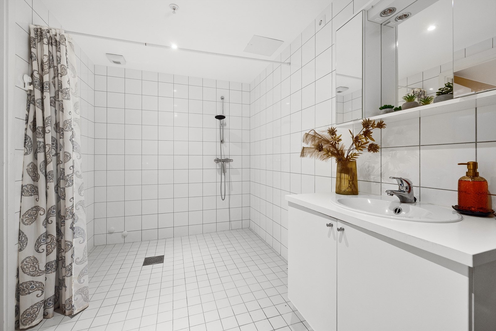 Badet inneholder romslig dusjsone med forheng, veggmontert toalett og baderomsinnredning med servantskap, servant med ett-greps blandebatteri og speil på vegg. Galleribilde