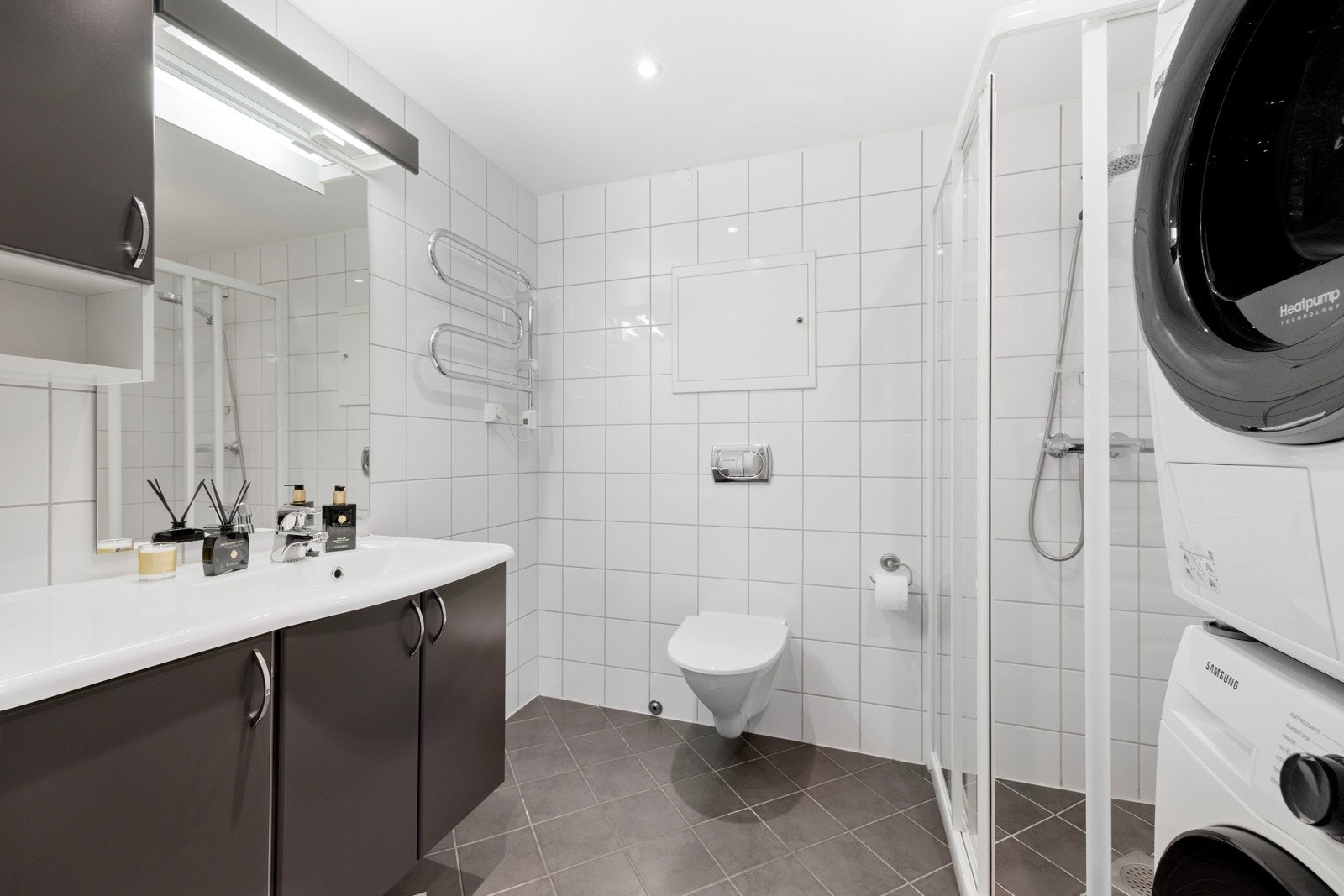 Lyst og delikat bad med fliser på gulv og vegger, varmekabler, innfelte spotter og ventilasjon. Badet har vegghengt toalett, dusjnisje, servant og opplegg til vaskemaskin - et praktisk og stilrent rom Galleribilde