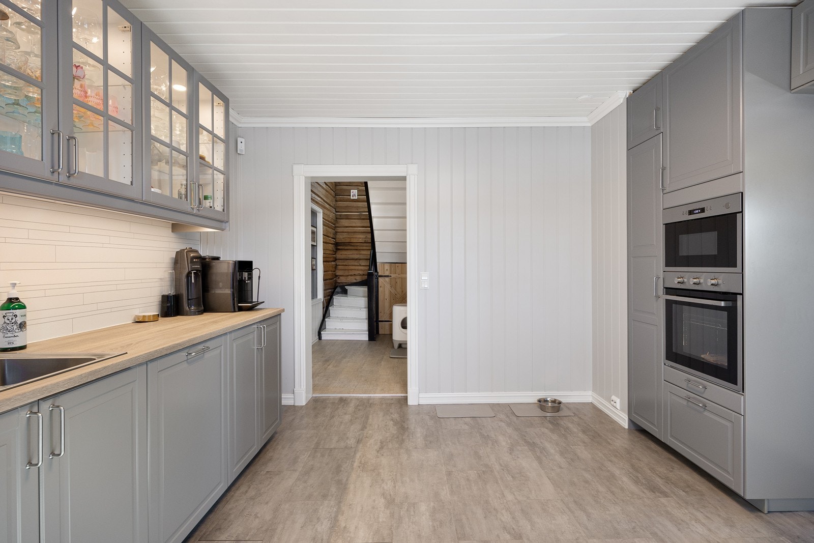 Kjøkkeninnredning med grå, profilerte fronter, flere vitrineskap med belysning og en lys laminatbenkeplate med nedfelt vaskekum i stål. Galleribilde