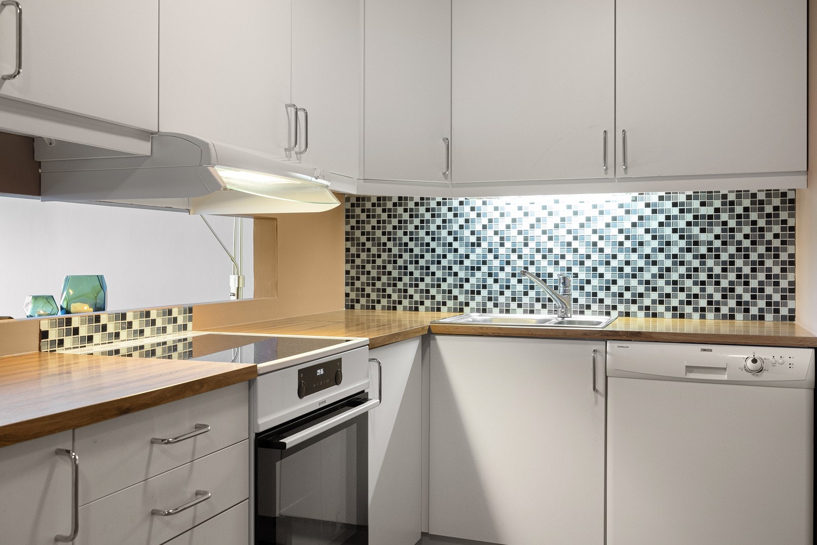 Kjøkkeninnredningen har hvite, glatte fronter, laminert benkeplate og flislagte overflater mellom overskap og kjøkkenbenken. Galleribilde