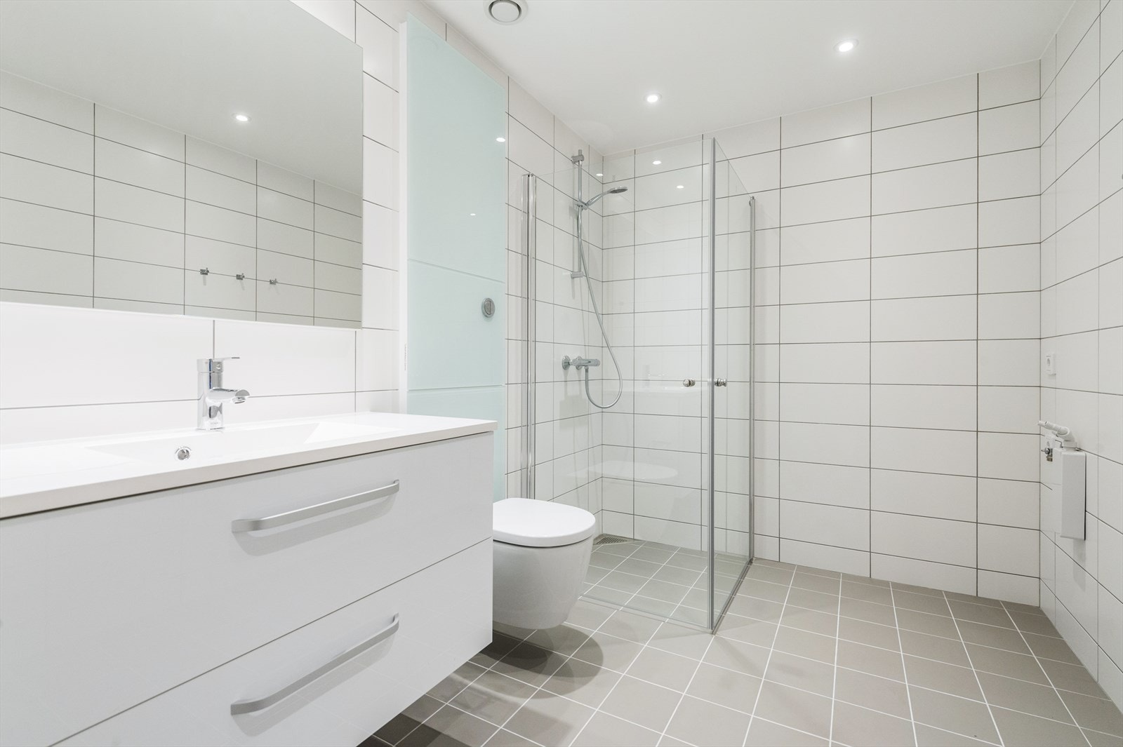 Moderne flislagt bad med dusjhjørne, vegghengt toalett og baderomsinnredning med speil og servant. Badet har ogsa opplegg til vaskemaskin og tørketrommel. (Alle bilder er hentet fra naboleilighet i samme etasje som er identisk.) Galleribilde