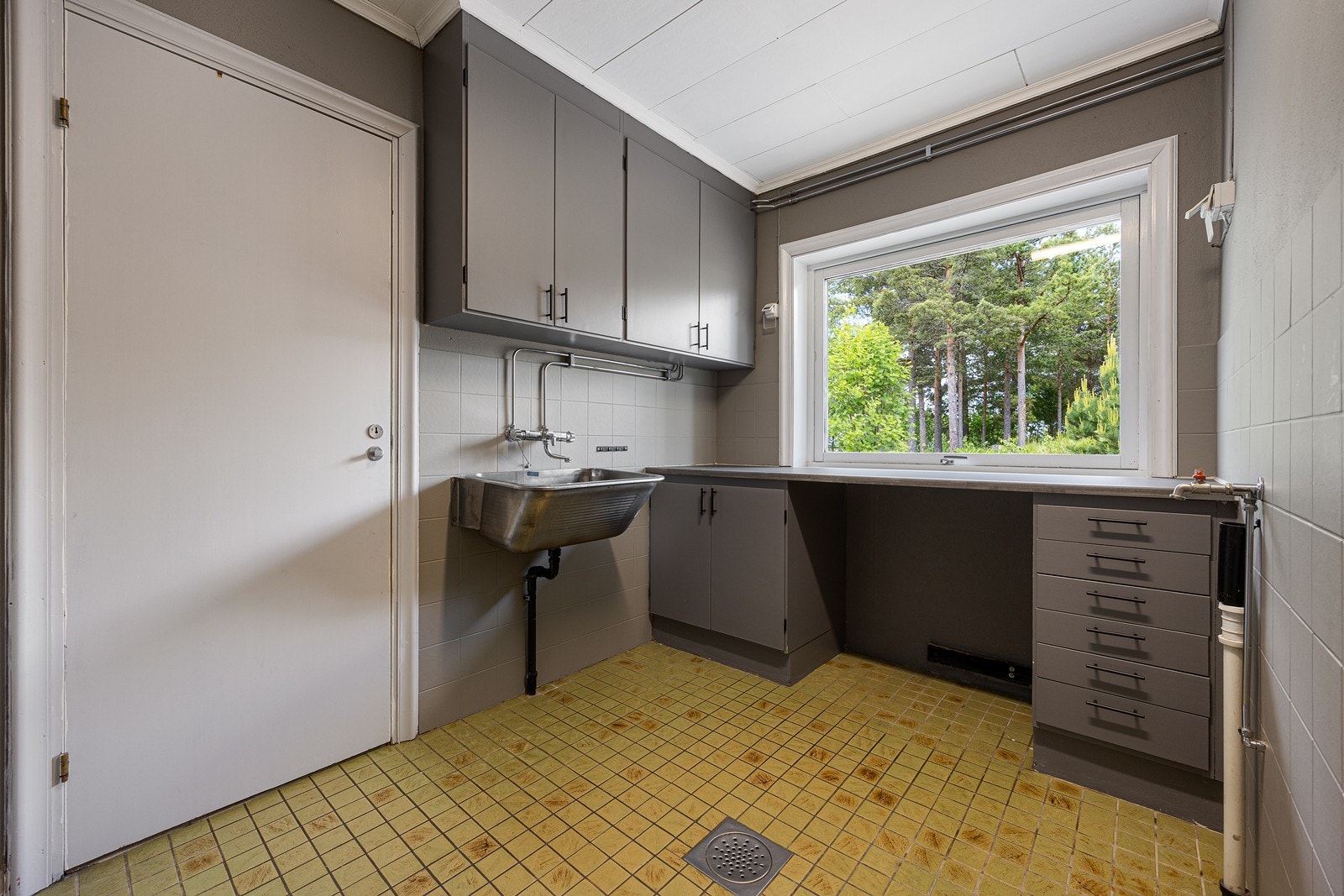 Praktisk vaskerom med gule fliser på gulv med vannbåren gulvvarme, gråmalte veggflater, et vindu og hvitmalt himling. Innredet med skyllekum, benkeinnredning og opplegg for vaskemaskin. Galleribilde