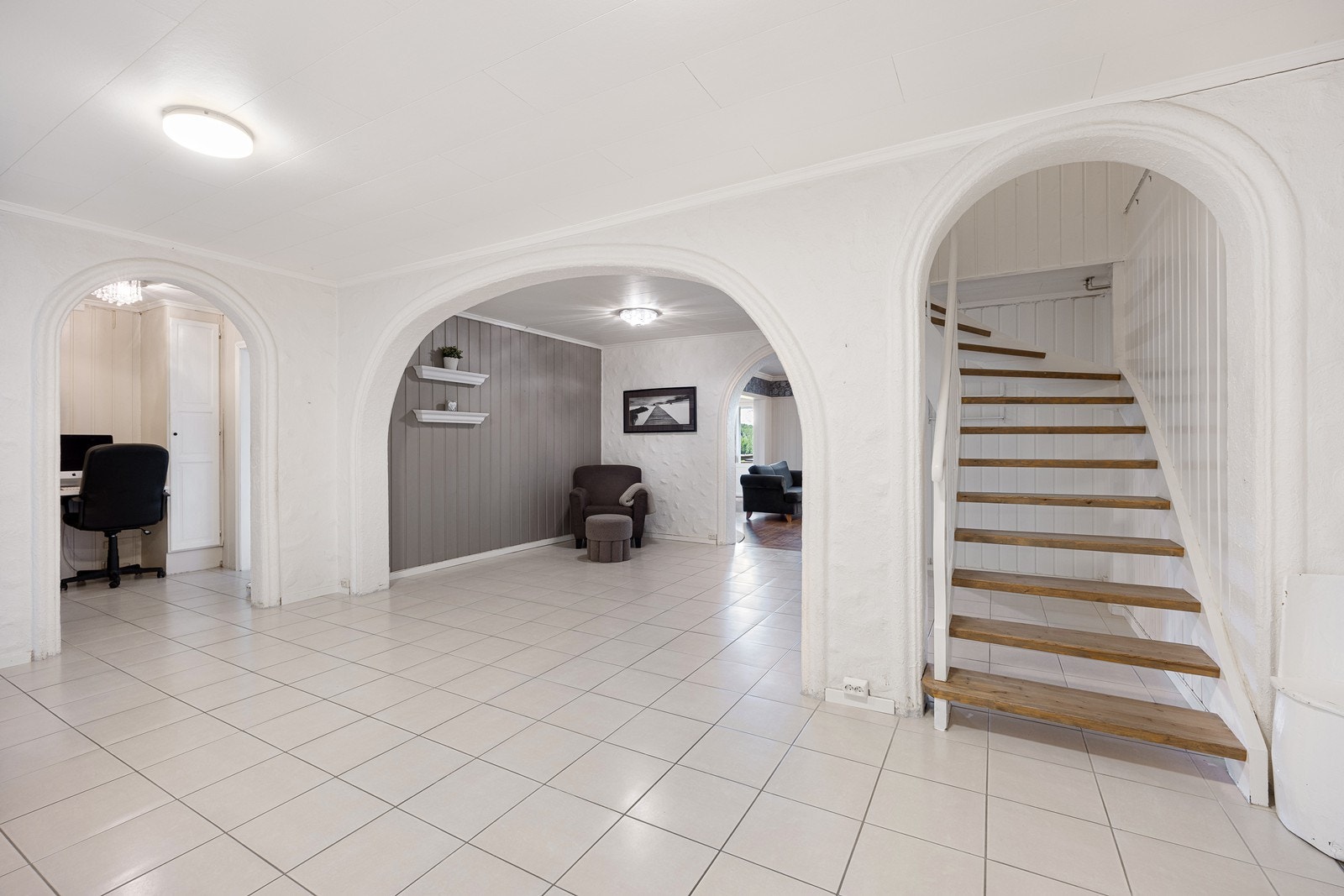 Stor hall med lyse gulvfliser, malte vegger i mur og panel, bueåpninger og en trapp med trehvite trinn opp til andre etasje. Galleribilde