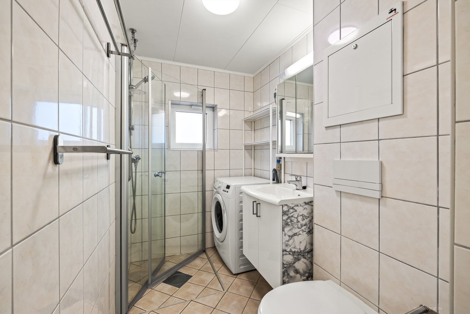 Det er fliser på vegger og gulv, varmekabler i gulv, vegghengt toalett, baderomsinnredning med servant, dusjhjørne med foldbare glassdører og opplegg til vaskemaskin. Galleribilde