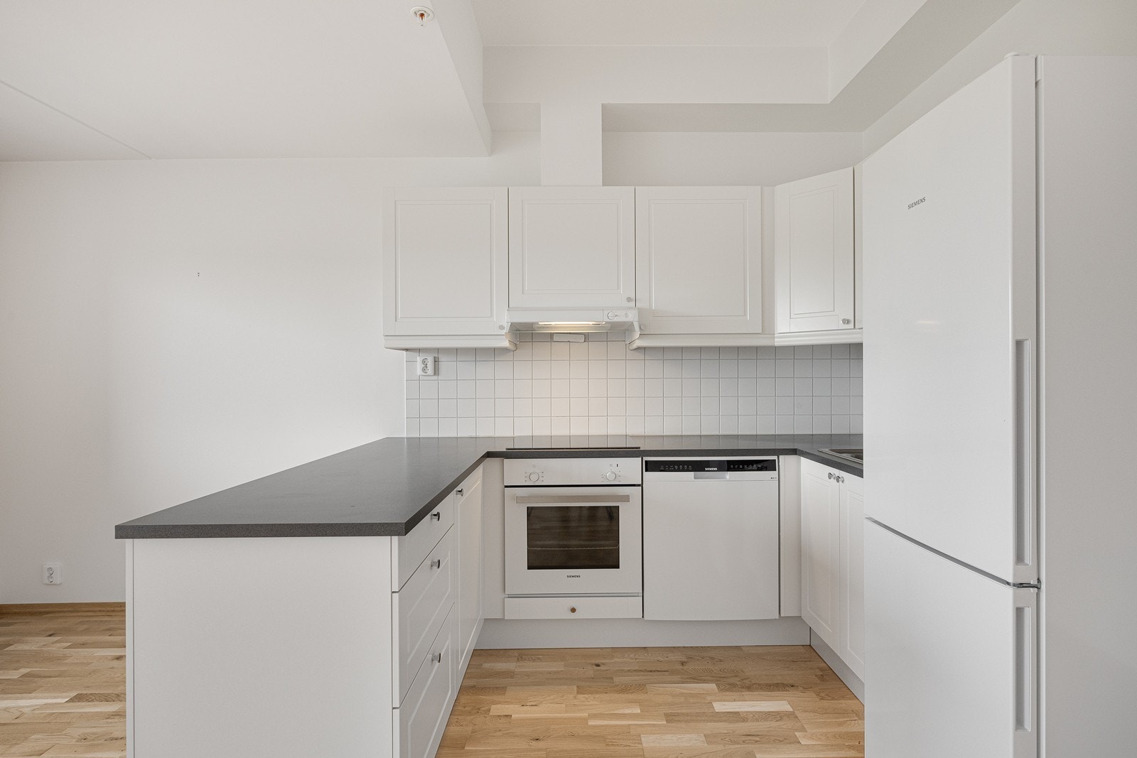 Det er fliser mellom kjøkkenbenken og overskapene. Kjøkkenet er utstyrt med integrert komfyr i benk og en nedfelt induksjon platetopp. Det er nisje for oppvaskmaskin i benk og avsatt plass til kjøl/frys. Galleribilde