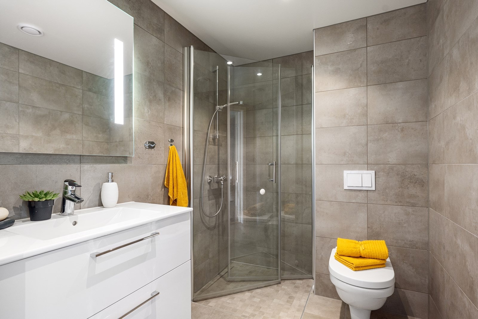 Badet i 2. etasje er stilfullt innredet med grå fliser og gulvvarme. Rommet er utstyrt med veggmontert toalett, dusjhjørne med glassdører og dusjbatteri fra Oras. Galleribilde