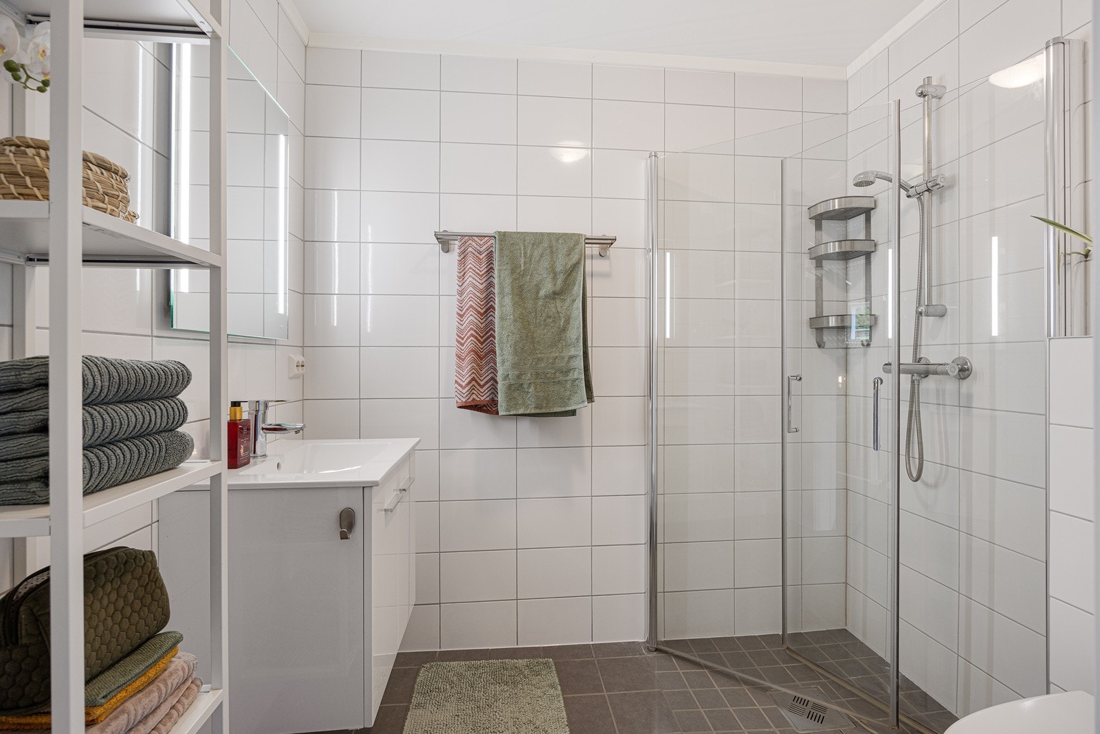 Badet gir et delikat inntrykk med veggmontert toalett og et dusjhjørne med innfellbare glassdører. Den hvite baderomsinnredningen har glatte skapfronter, heldekkende servant og speil med integrerte belysning. Galleribilde