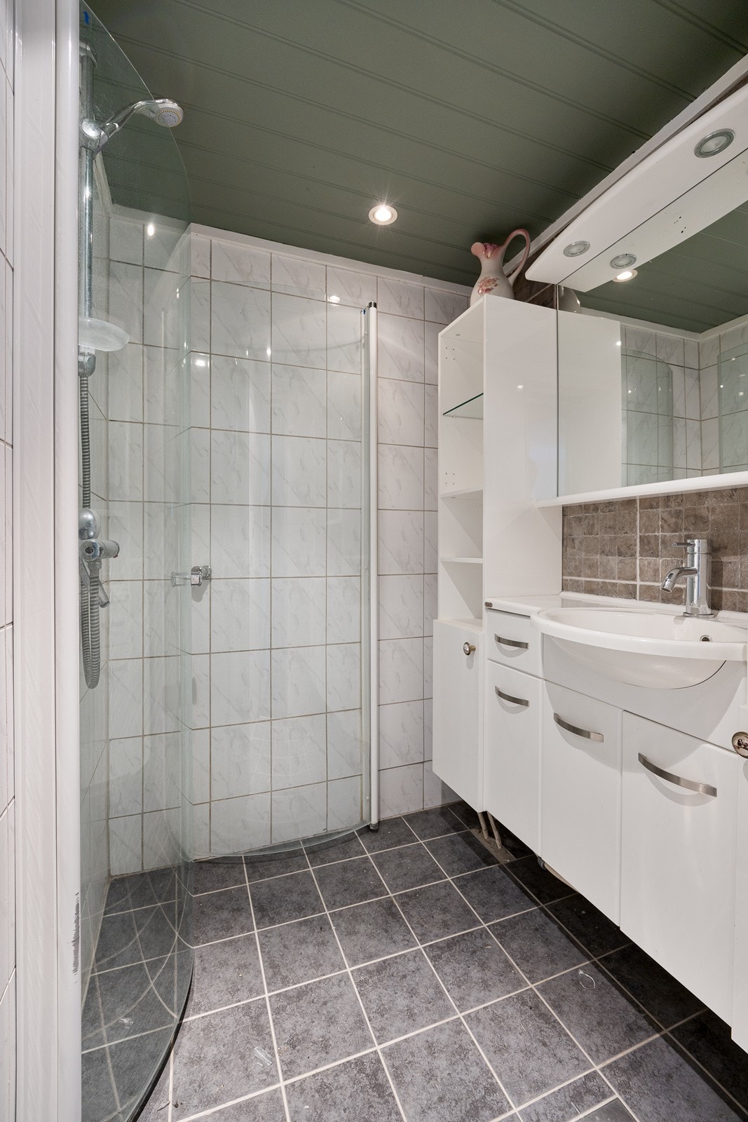 Badet i underetasjen har grå gulvfliser med gulvvarme, lyse veggfliser og mørkmalt panelhimling med downlights. Galleribilde