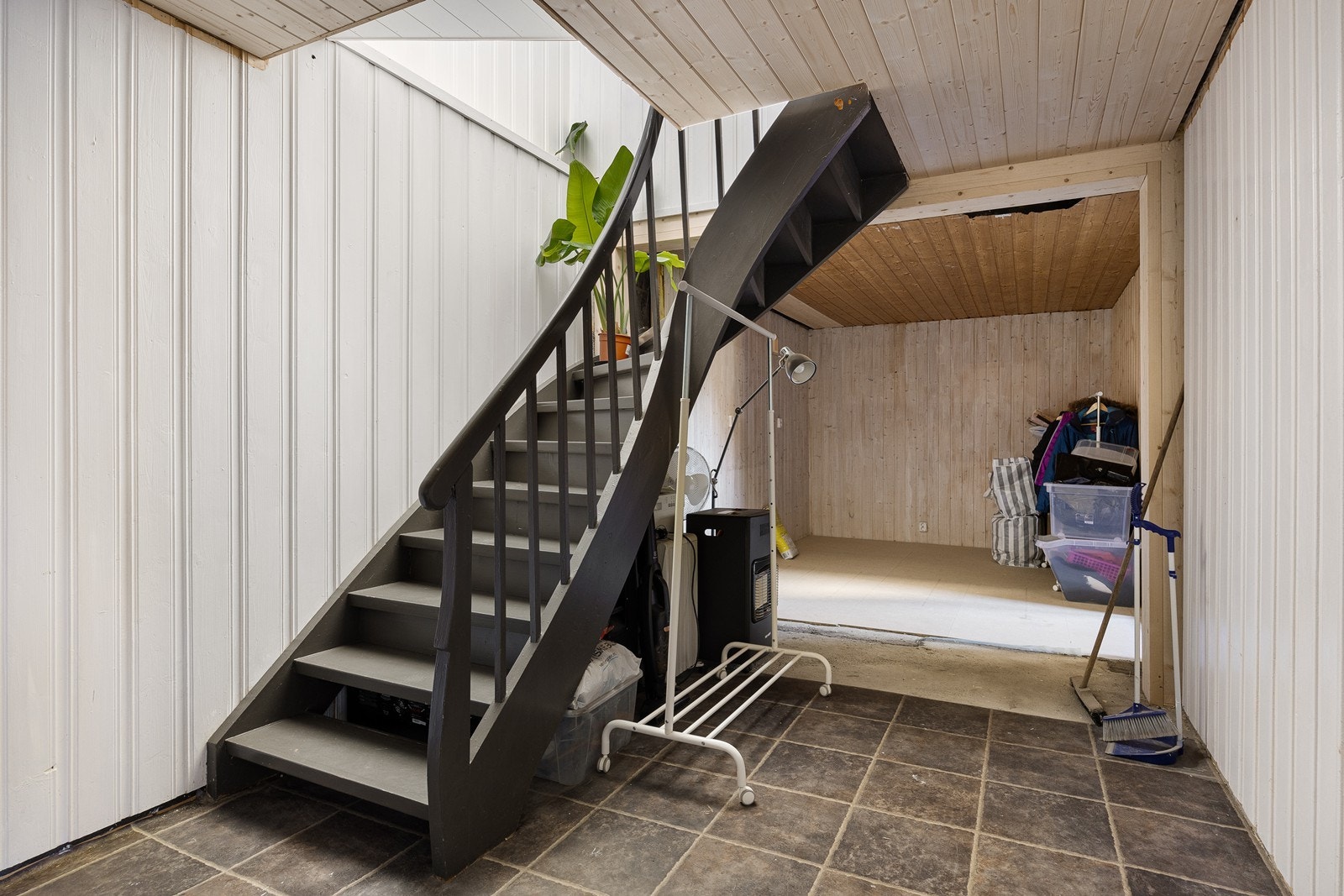 Fra entreen fører en mørkmalt trapp ned til underetasjen, med flislagt gang, paneltak og hvitmalte panelvegger. Galleribilde