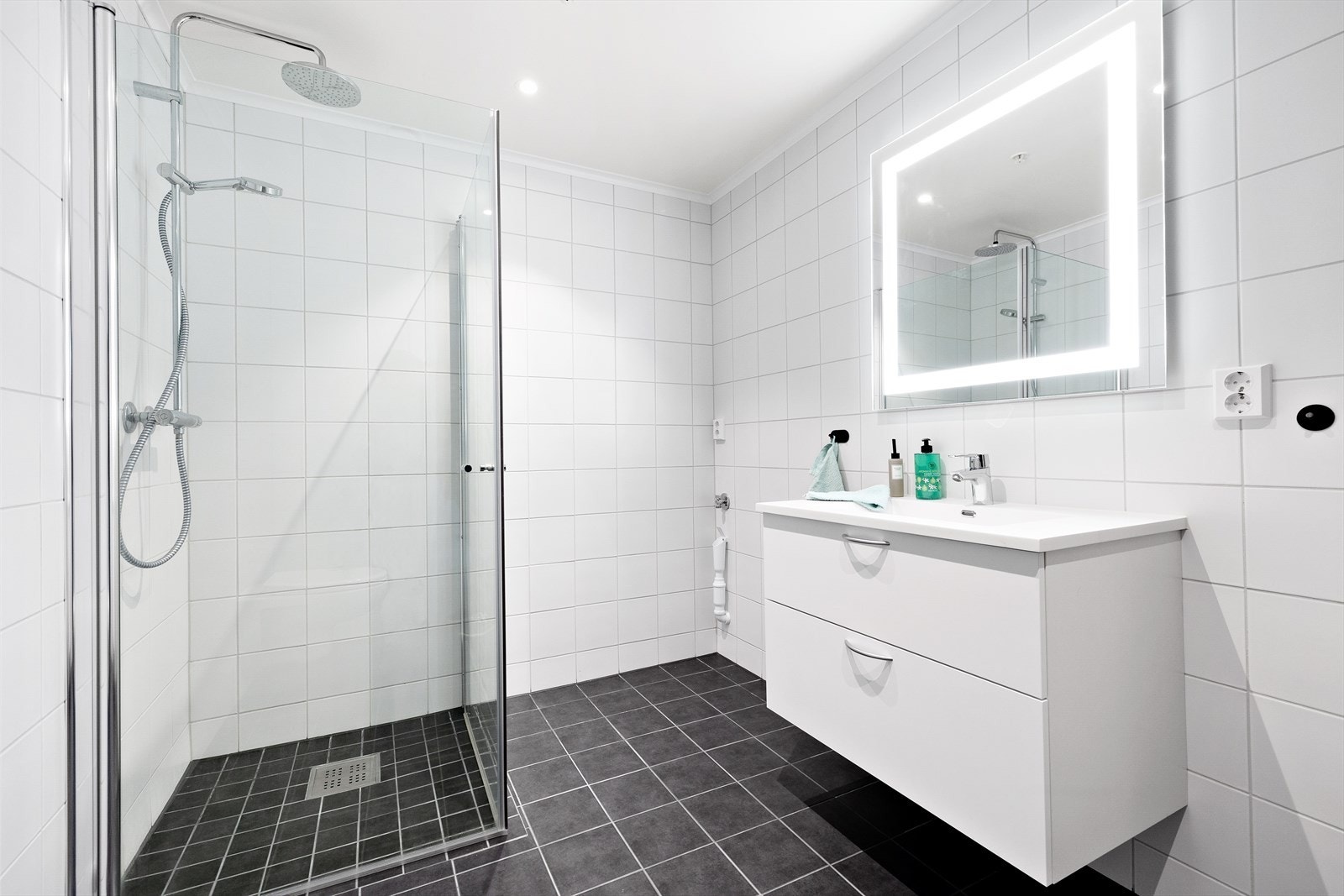 Lekkert og stilrent bad med fliser på gulv og vegger. Varmekabler i gulv. Inneholder servantinnredning, vegghengt toalett, dusj og opplegg for vaskemaskin og tørketrommel. Galleribilde