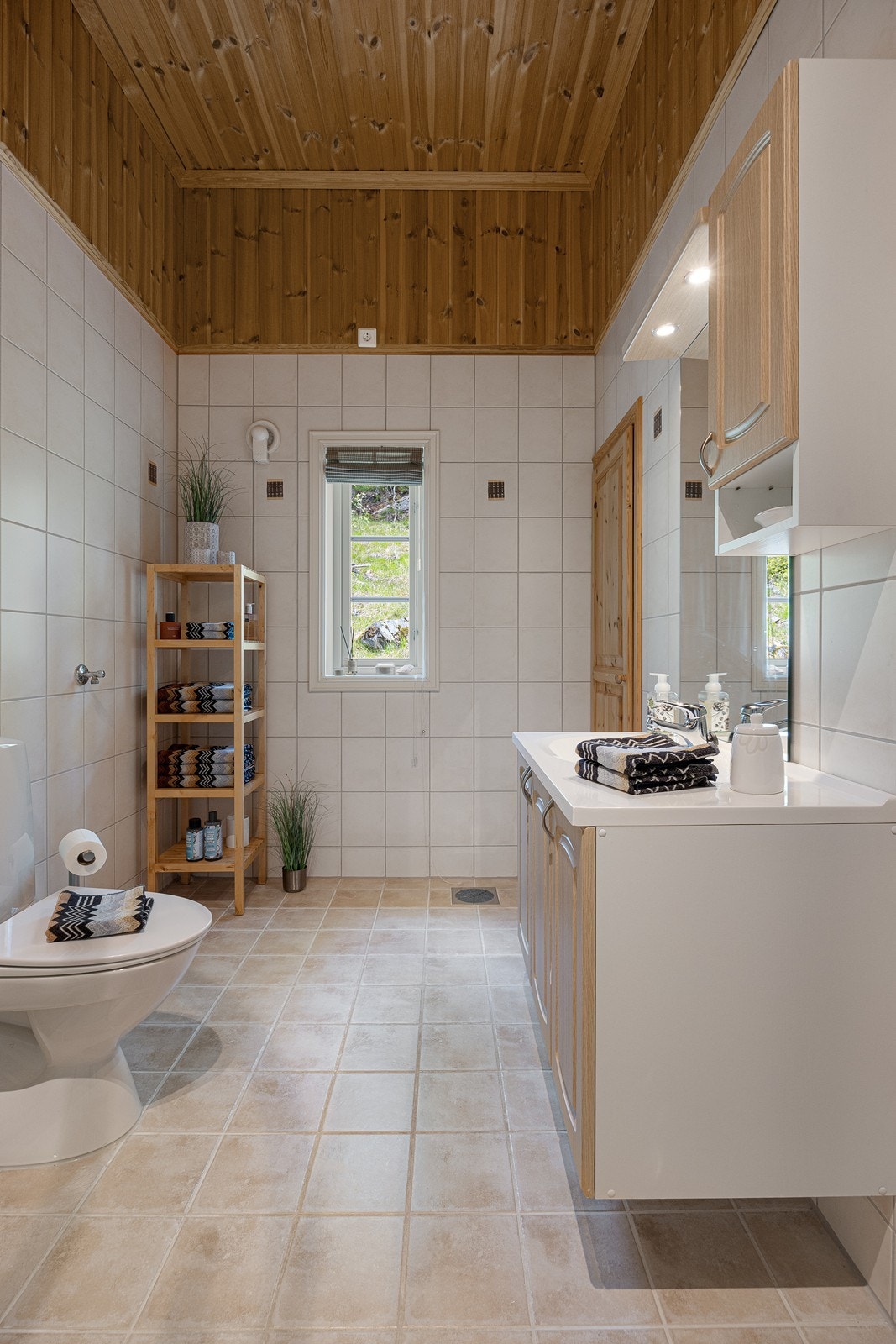 Baderomsinnredningen har lyse, profilerte fronter og består av en heldekkende servant på en skapseksjon, et veggskap og et stort speil med belysning. Videre har badet et toalett, opplegg for vaskemaskin og et dusjhjørne med skyvedør i glass. Galleribilde