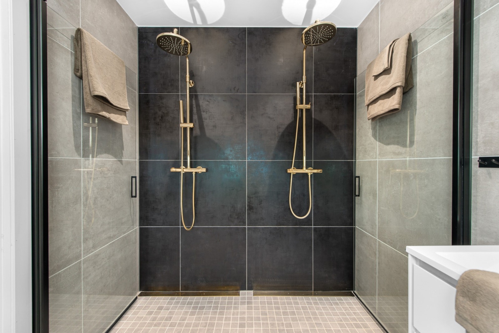 Her er det en delikat dusjsone med 2 regnfallsdusjer i gullfarge, 60x60 fliser på vegger og innfellbare dusjdører i glass. Galleribilde