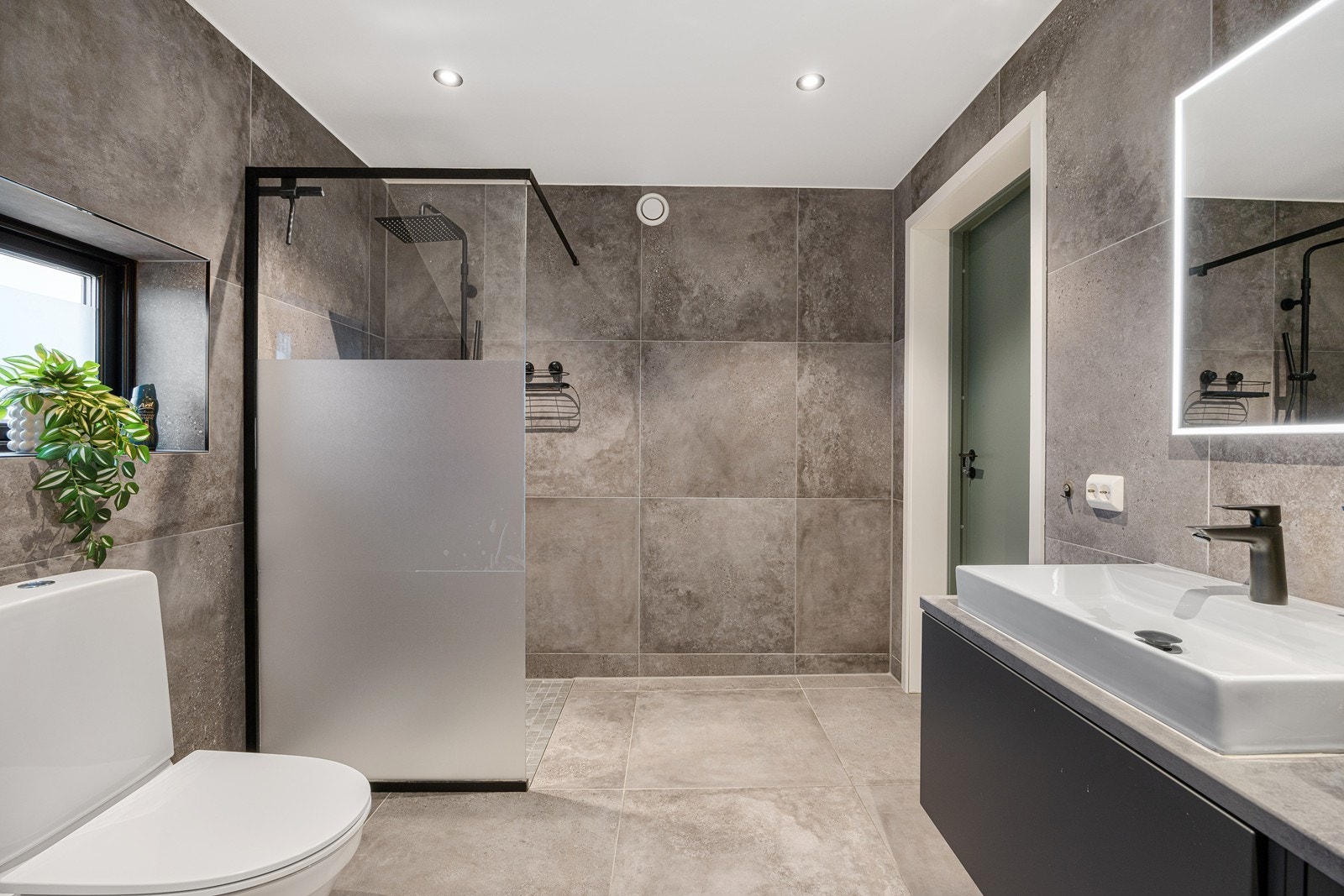 Badet oppe har et industrielt preg på flisene; store og stilige i ulike gråfarger. Dusjhjørnet har glassvegg, mosaikkfliser og sort regnfallsdusj. Galleribilde