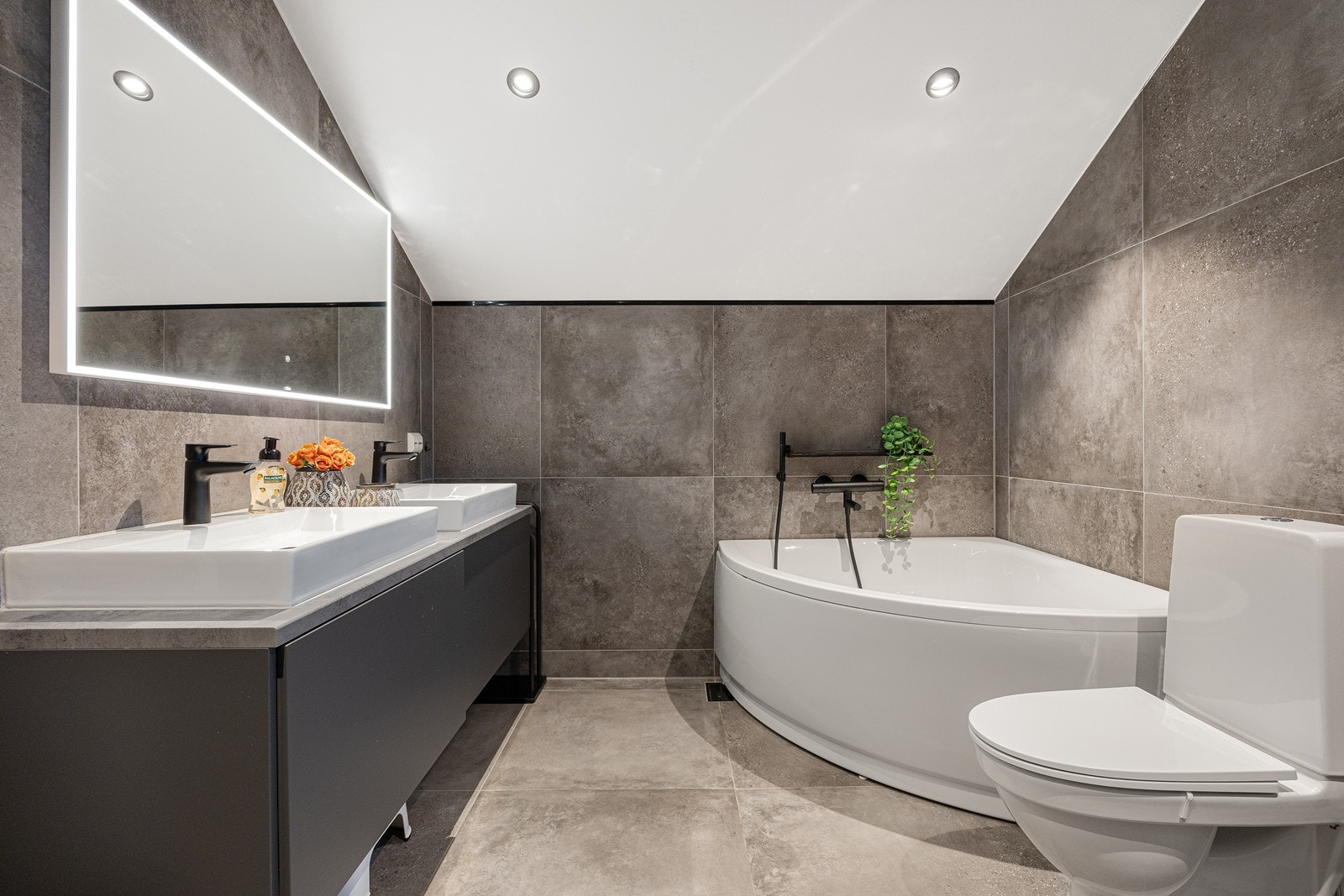 Varme er lagt i gulvet, og badet er innredet med toalett og downlights i taket. Baderomsinnredningen består av et stort speil med integrert belysning og en mørk skapseksjon med to toppmonterte servanter. Galleribilde