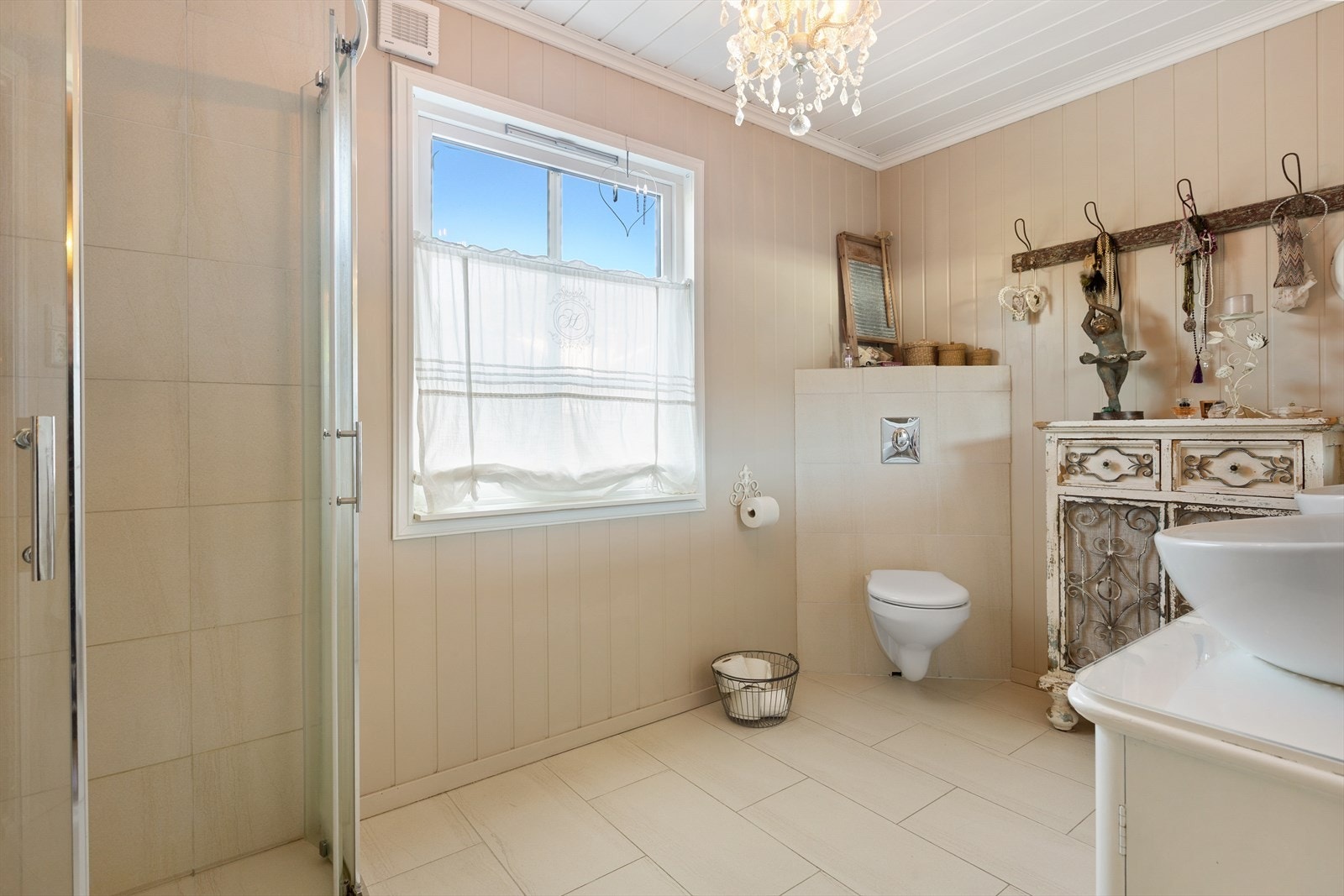 Heltre skapinnredning med to porselensservanter. Flislagt dusjhjørne med glassdører og veggehengt toalett med innfliset sisterne. Det er bryterstyrt vifte i vegg. Galleribilde