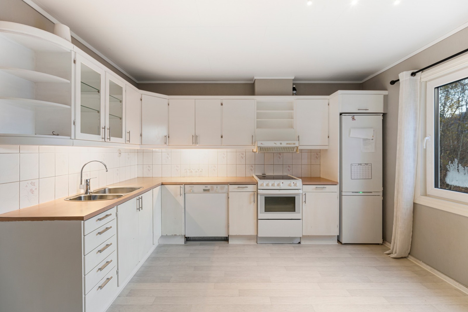 Kjøkkenet har en L-formet innredning bestående av benkeskap og overskap, og opplegg for frittstående hvitevarer Galleribilde