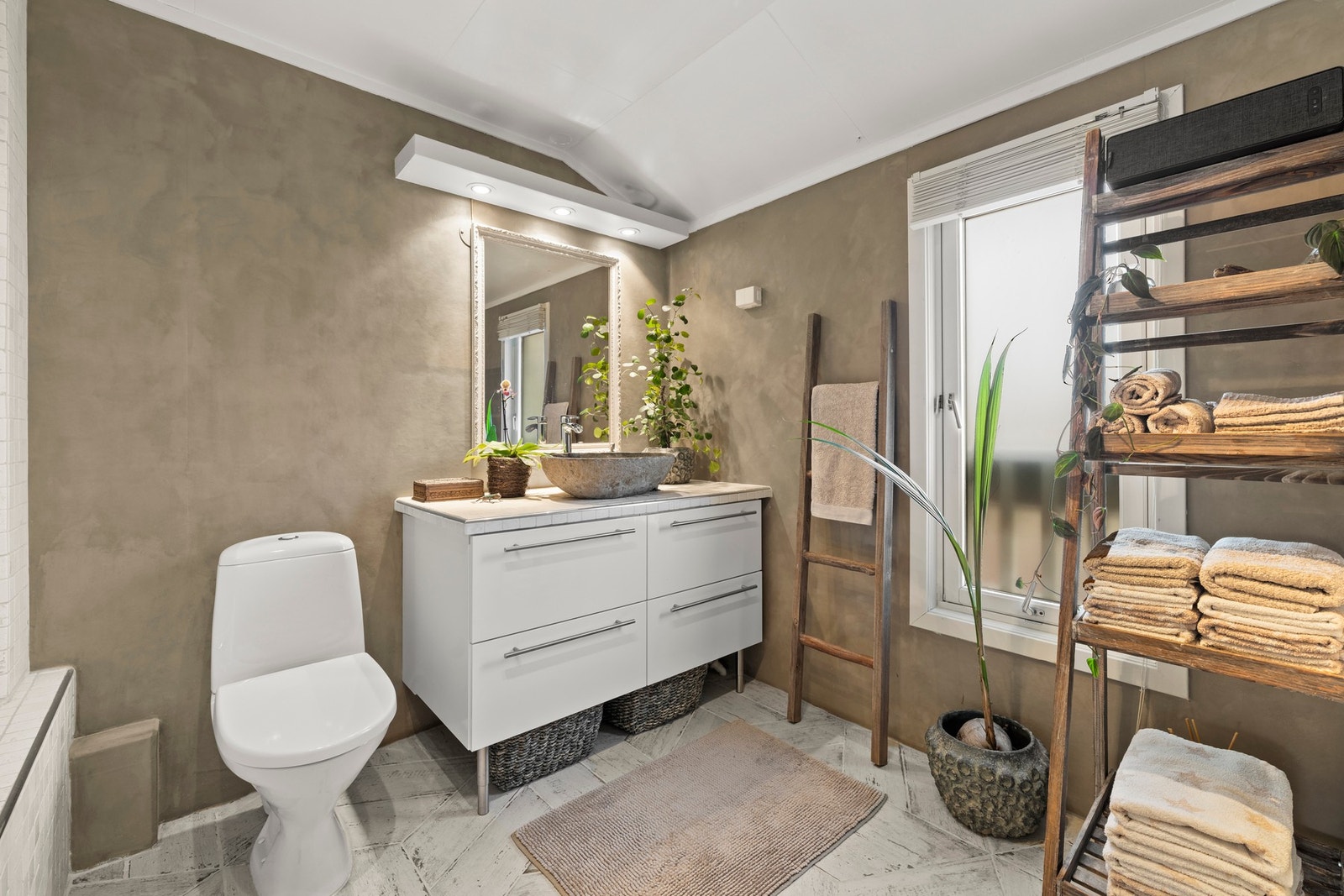Badet fremstår stilrent og funksjonelt, med flislagte gulv- og veggflater kombinert med moderne mikrosementdetaljer. Varmekabler i gulvet gir god komfort, og belysning i både tak og over speil sørger for et lyst og behagelig rom. Det er mekanisk avtrekk v Galleribilde