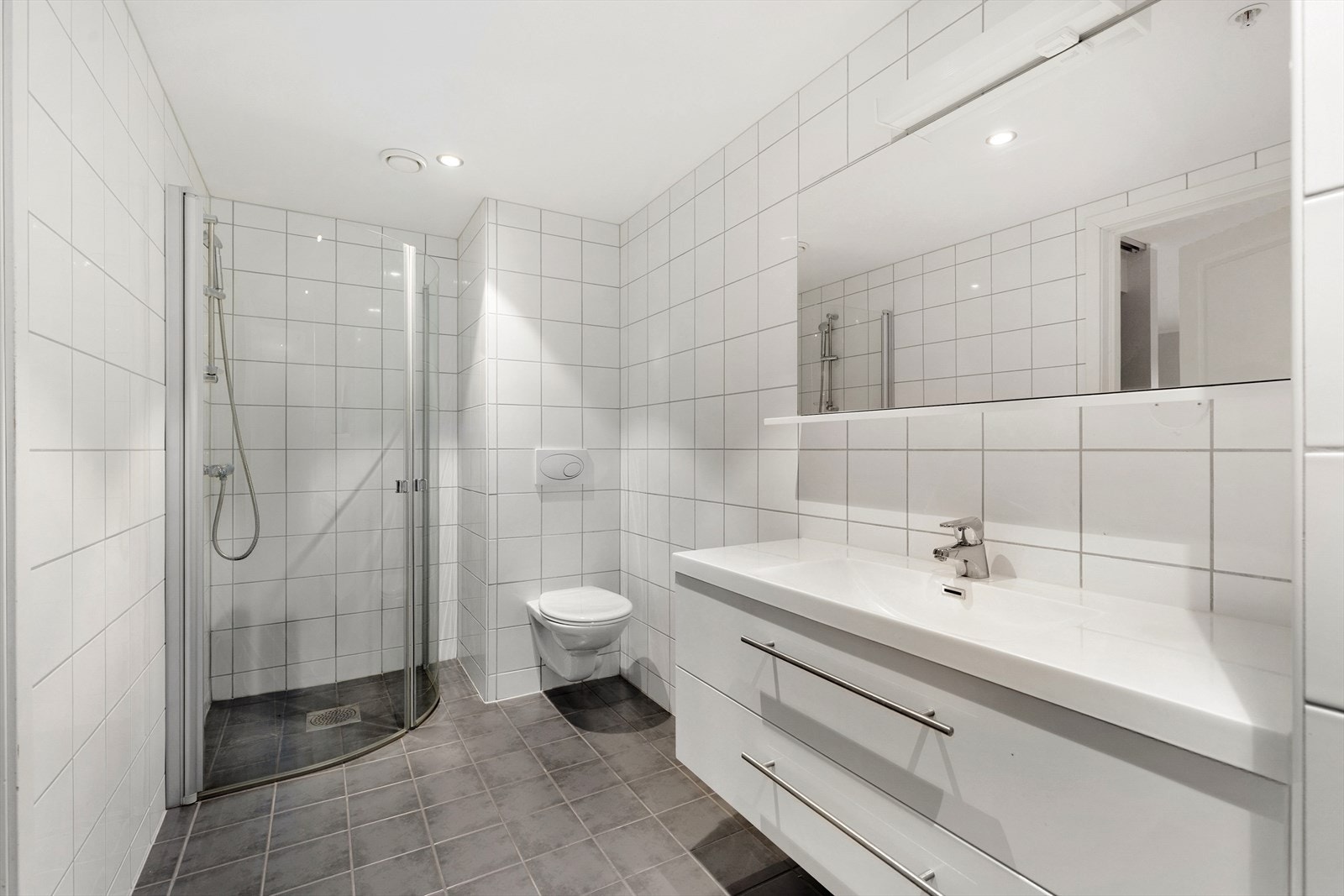 Lyst, flislagt bad med varmekabler i gulv og innfelte spotter i tak, samt veggmontert toalett og dusjnisje med dører i herdet glass. Galleribilde