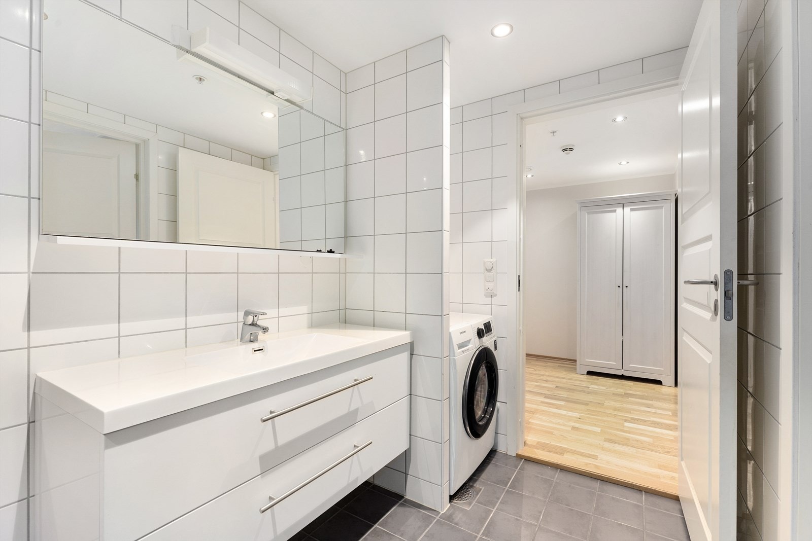 Bred servantinnredning med skuffer og heldekkende servant, samt speil på vegg. Egen nisje med opplegg for vaskemaskin. Galleribilde