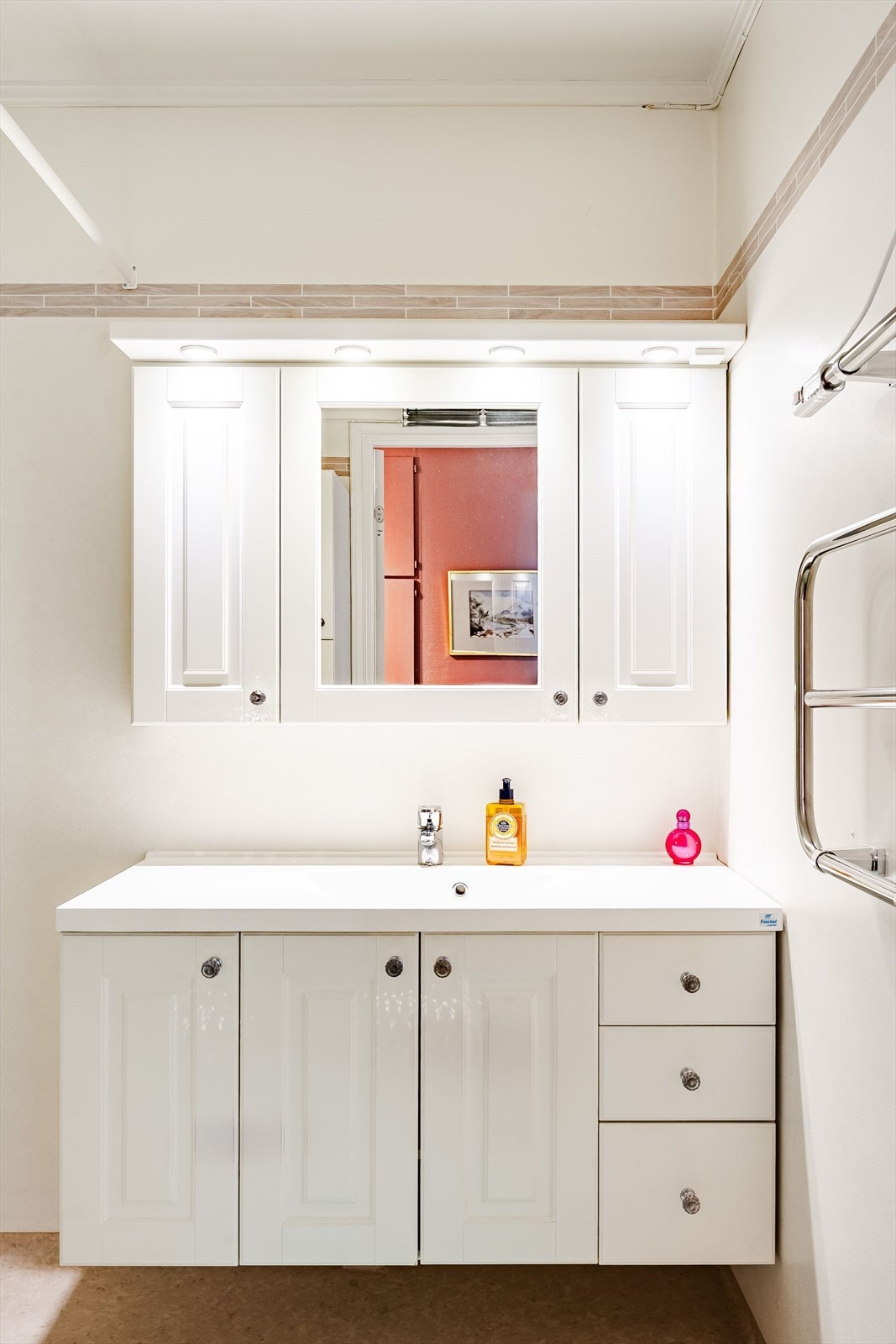 Tidløs baderomsinnredning med lyse profilerte fronter og heldekkende servant, samt vegghengte skap med belysning over. Baderommet er også utstyrt med håndkletørker, dusjhjørne med forheng og opplegg for vaskemaskin. Galleribilde