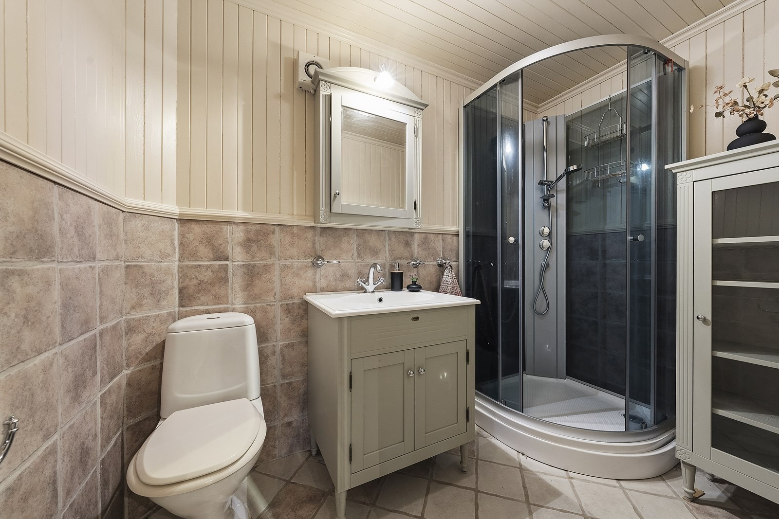 Baderom med flislagt gulv med varmekabler. Badet er utstyrt med servant over skap, speilskap, toalett og dusjkabinett Galleribilde
