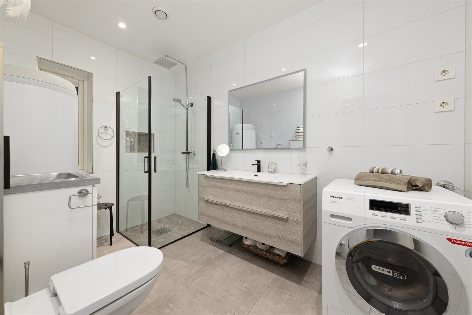 Bad/vaskerom med dusjhjørne, vegghengt toalett, opplegg for vaskemaskin og moderne baderomsinnredning. Galleribilde