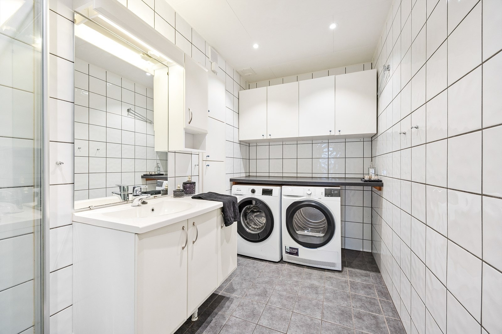 Badet er utstyrt med dusjhjørne med innfellbare glassvegger og servantinnredning med under- og overskap, samt opplegg og plass til både vaskemaskin og tørketrommel under praktisk brettebenk Galleribilde