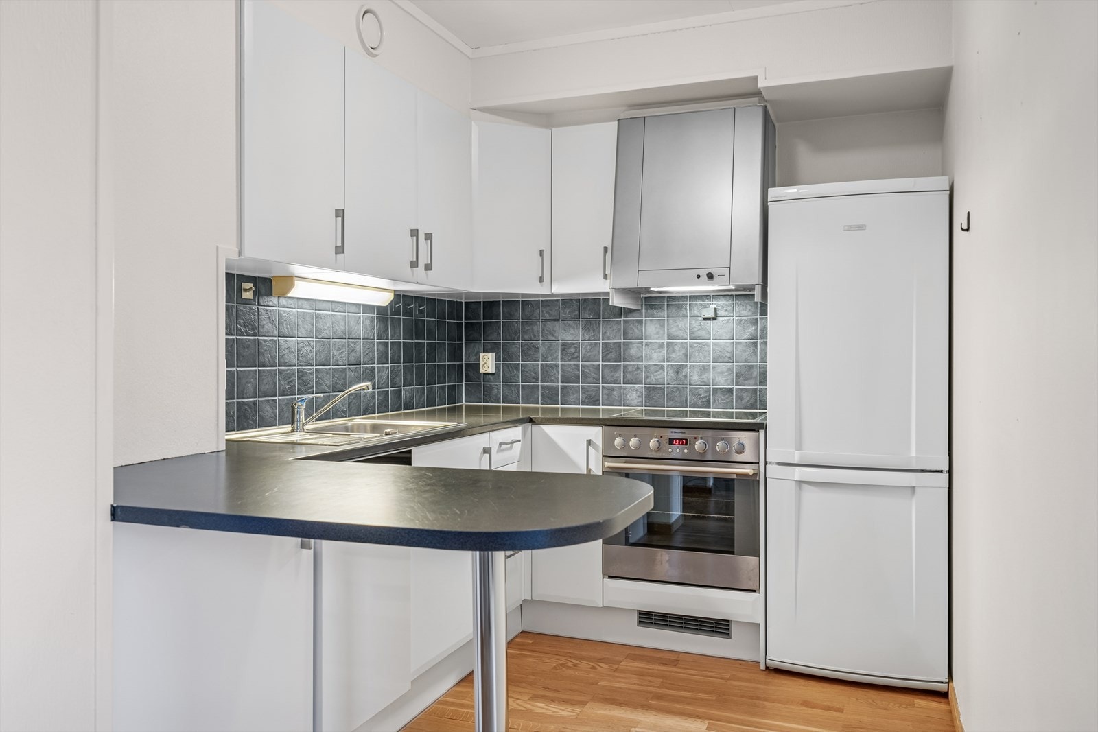 Kjøkkenet har innredning med malte slette fronter, laminert benkeplate med nedfelt stålvask og fliser på vegg over benk Galleribilde