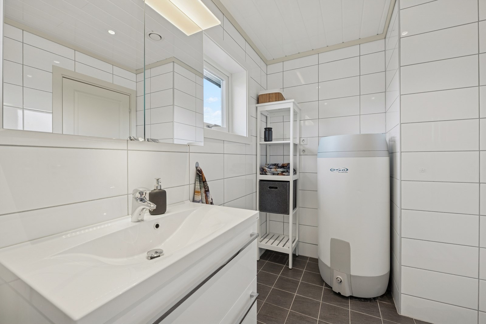 Det er en fin overgang mellom gulv- og veggflisene. Rommet er innredet med romslig baderomsinnredning med skuffer, dusjsone med glassdører, vegghengt toalett, opplegg for vaskemaskin og belyst speilskap. Galleribilde