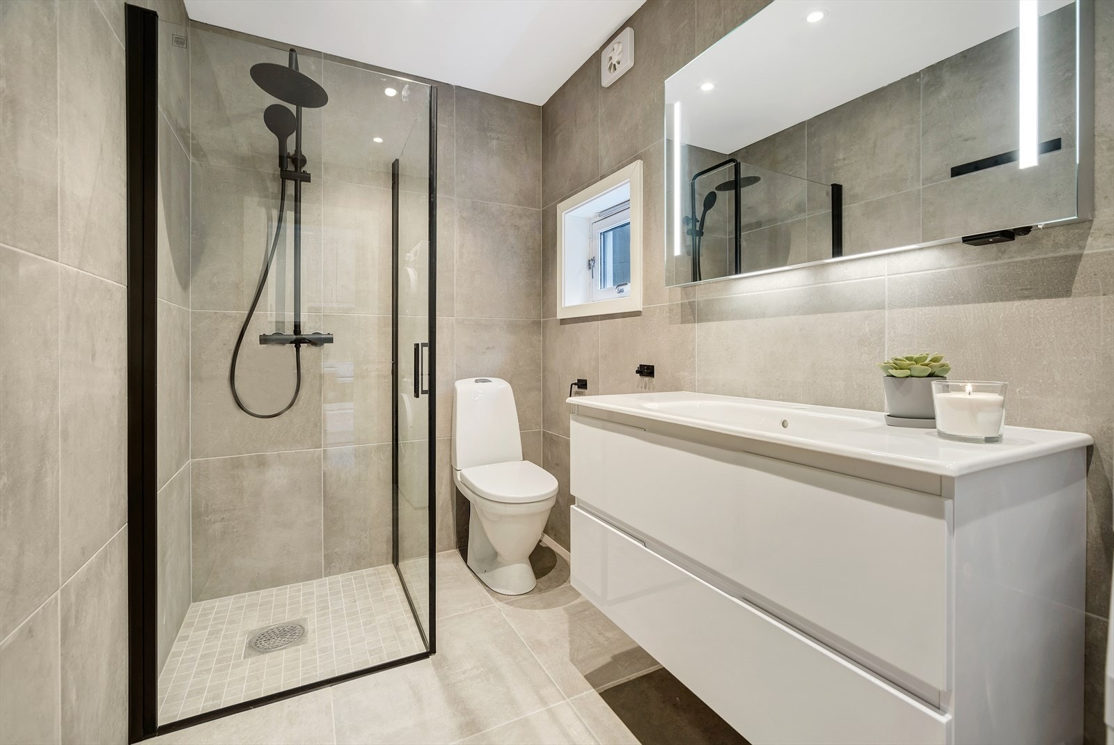 Et stilrent bad i moderne standard med flislagte vegger- og gulv. Det er innredet med opplegg for vaskemaskin, servant med underskap, toalett og hjørnedusj med innfellbare glassdører. Galleribilde