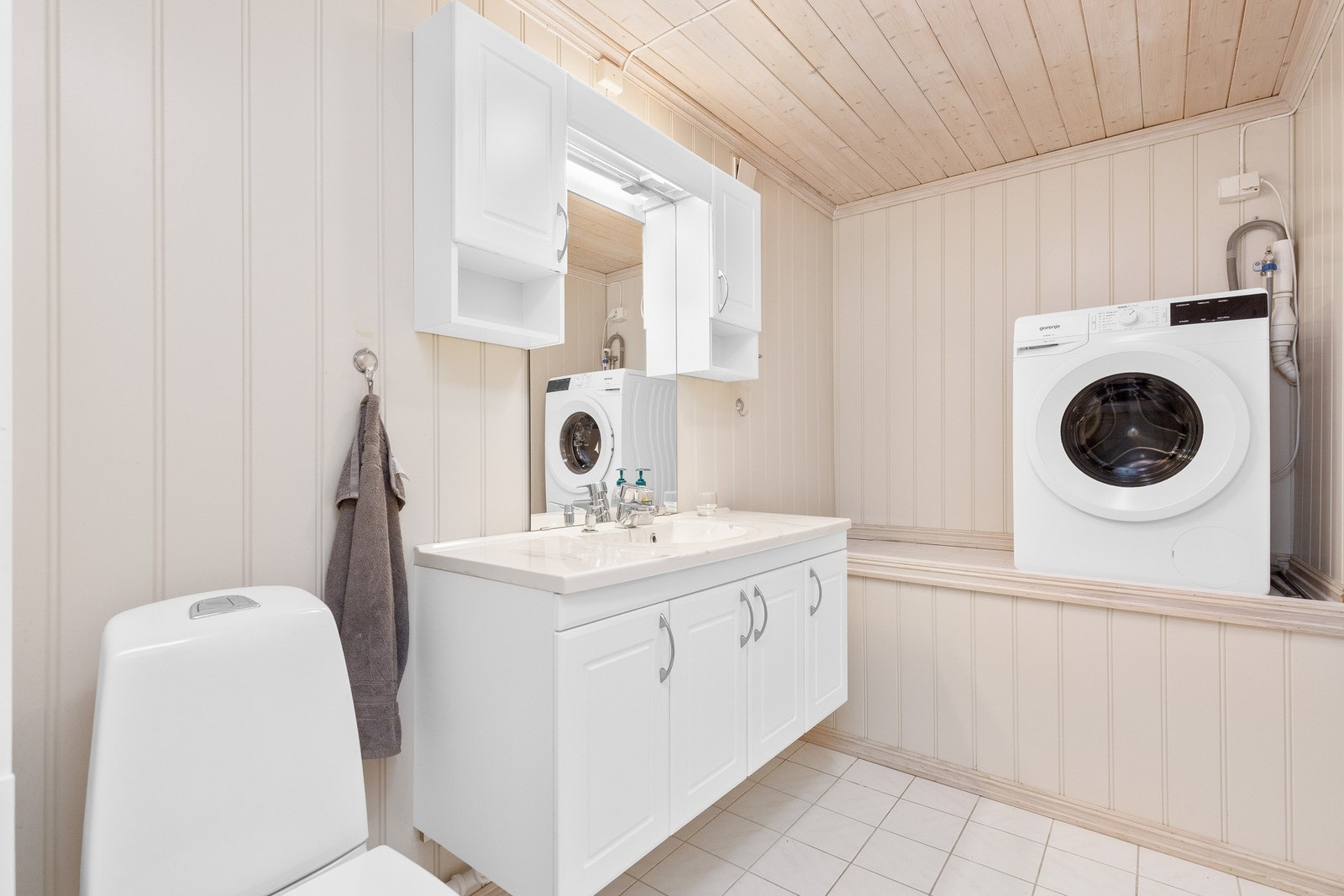 Lyst bad med panelvegger, flislagt gulv, opplegg for vaskemaskin, toalett og stort speilskap over servant. Galleribilde