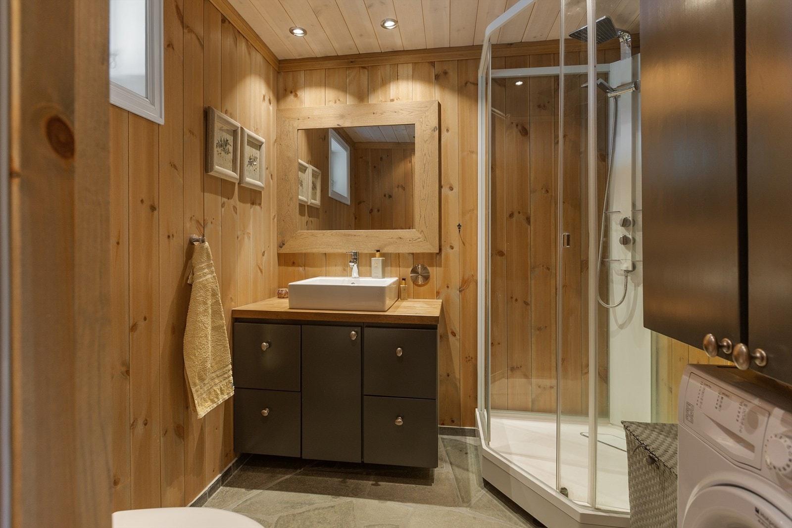 På badet er det gulv belagt med skifer og varmekabler. Stilig baderomsinnredning i heltre med underskap, servant, heltre benkeplate i eik og speil med eikeomramming. På badet er det også toalett, dusjkabinett og opplegg til vaskemaskin. Galleribilde