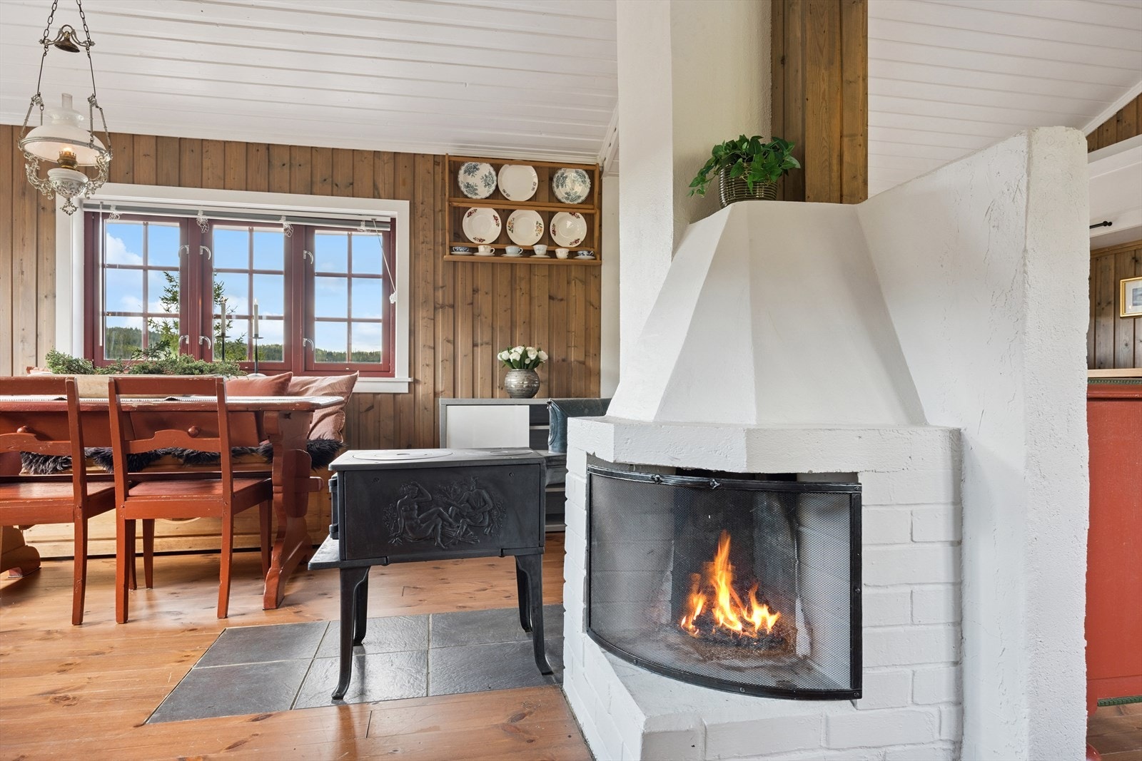 En åpen peis med hvitmalt brannmur både pynter opp og deler rommet. I tillegg gir vedovnen ved siden av peisen lun varme til hele hytta. Galleribilde