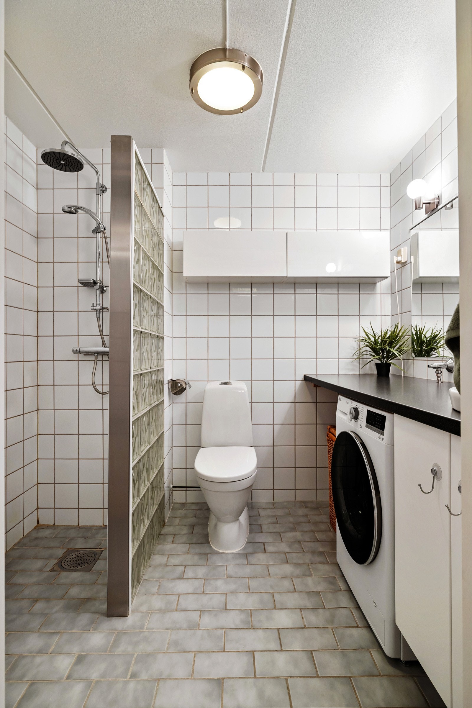 Badet er helfliset med lyse veggfliser og grå gulvfliser. Dusjsonen er avgrenset med glassbyggerstein og har både dusjhode og regnfalldusj. Rommet er innredet med servant på benk med underskap, speil med belysning over Galleribilde