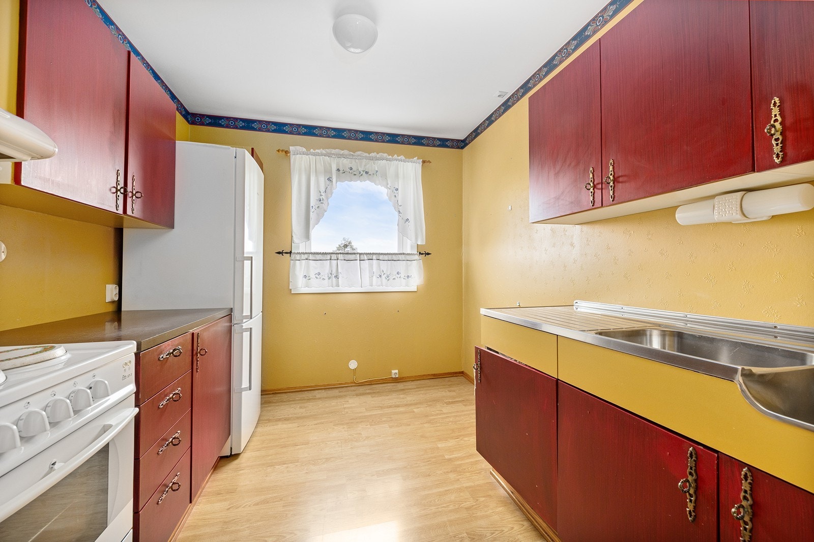 Kjøkkenet har innredning med glatte fronter. Benkeplaten er av laminat. Det er kjøl/fryseskap og komfyr. Stålbenk med kum og skyllekum. Galleribilde