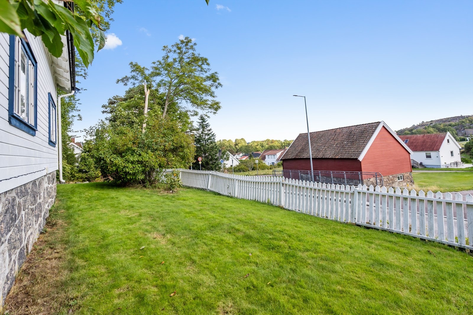 Tomten grenser til både vei og nabolag, men har en skjermet beliggenhet bak hekk og vegetasjon. Eiendommen har hvitt stakittgjerde mot veisiden og forstøtningsmur i naturstein. Galleribilde