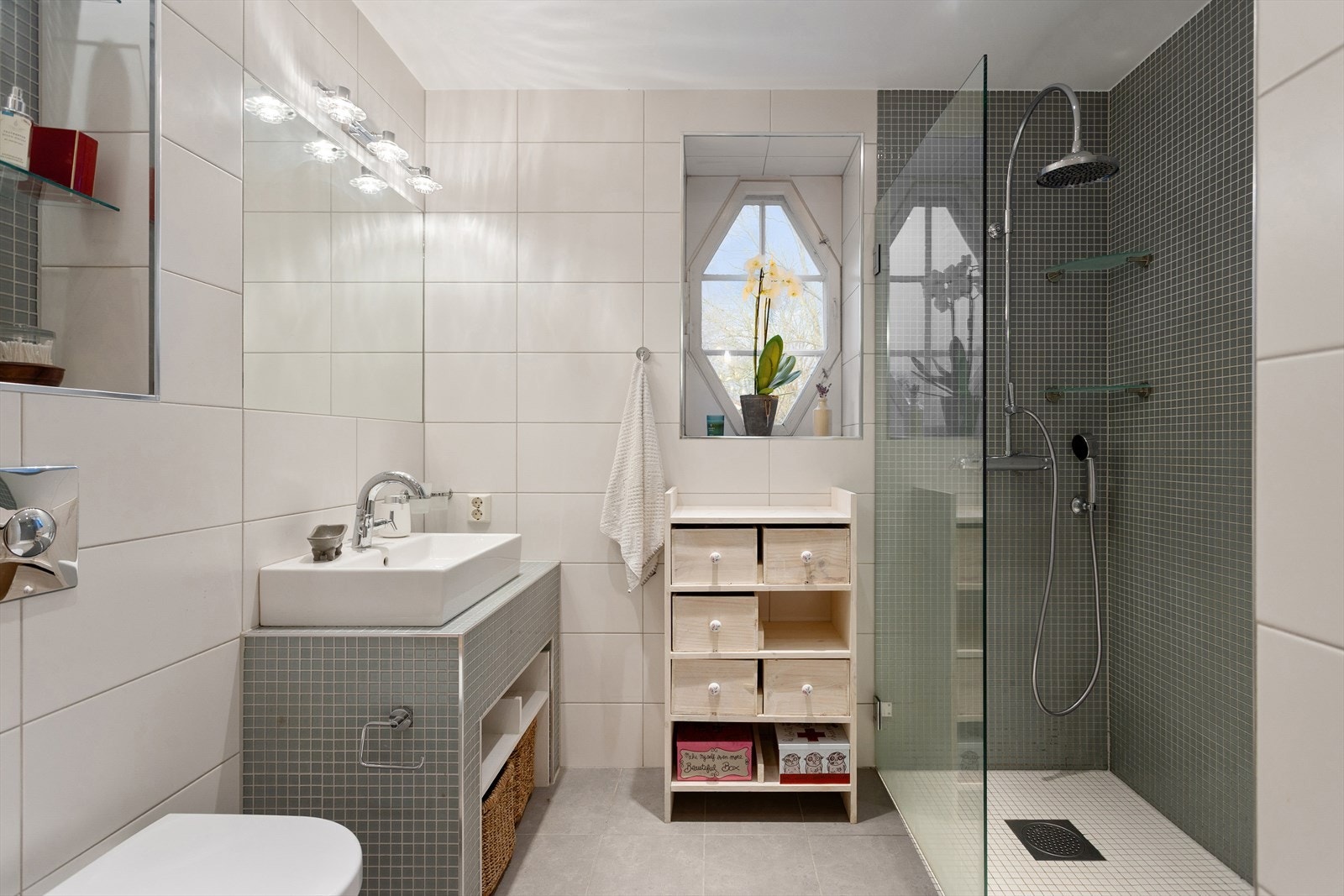 Badet er utstyrt med vegghengt wc, servant på mosaikk flislagt benk, dusj i hjørne med glassvegg/dør og elektrisk håndkletørker. Galleribilde