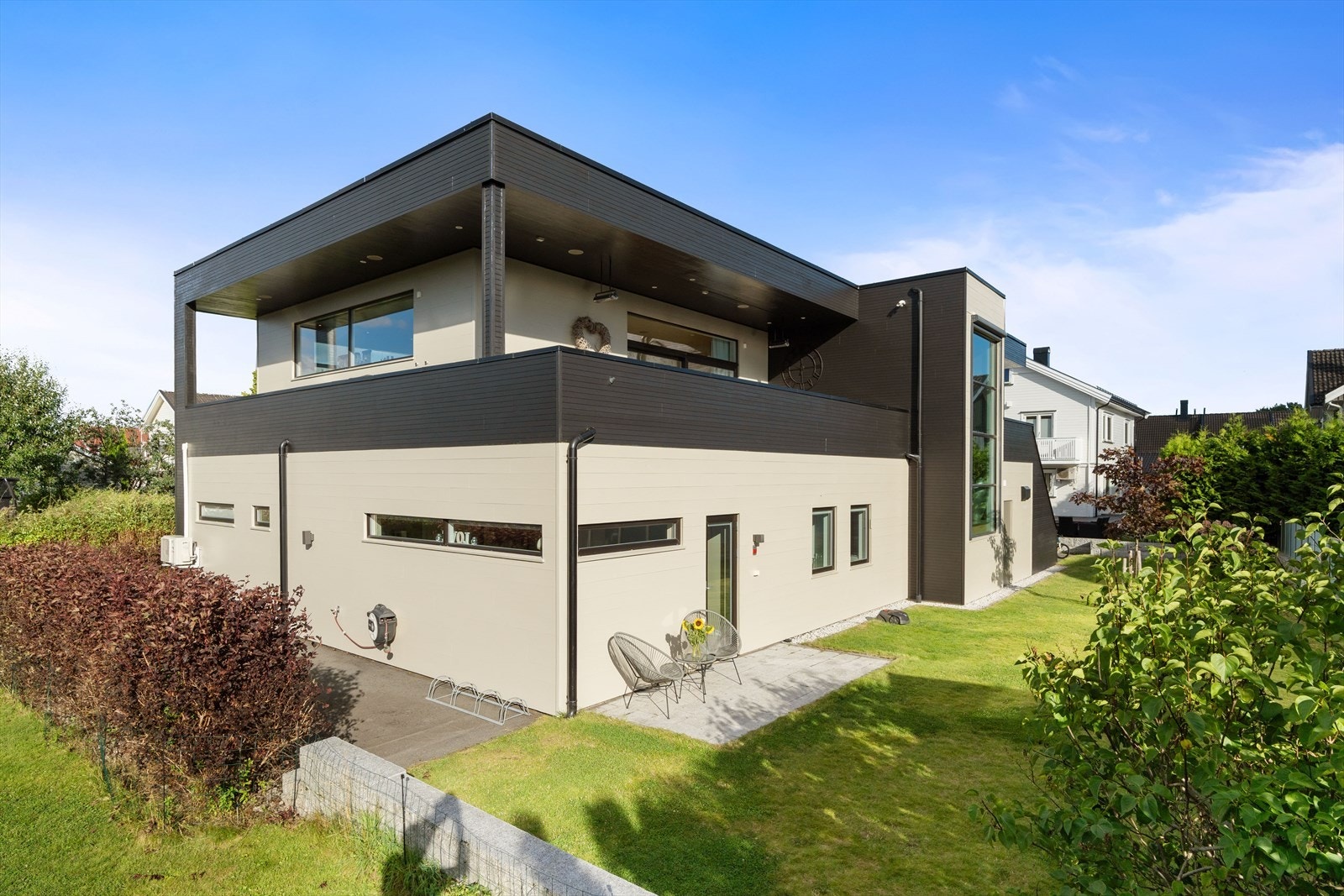 Boligen har et stilrent og minimalistisk uttrykk, med en fasade som kombinerer lyse og mørke farger - lyse vegger i øvre etasje og mørkere paneler i nedre del - som gir et elegant og tidløst preg. Galleribilde