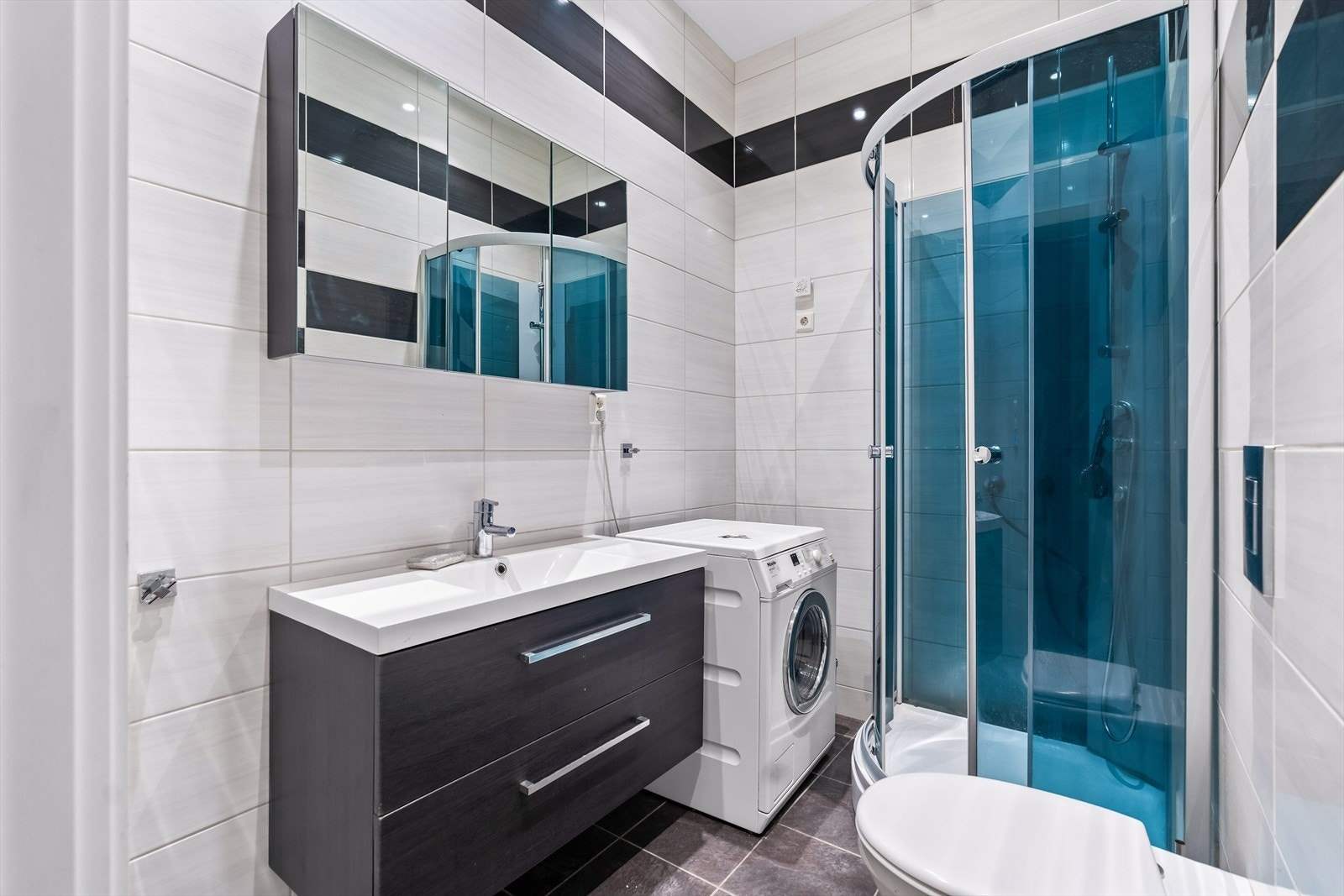 Badet har flislagte overflater. Rommet er videre utstyrt med baderomsinnnredning med servant og ett-grepsblandebatteri, vegghengt toalett og dusjhjørne med glassdører. I tillegg har badet opplegg for vaskemaskin. Galleribilde