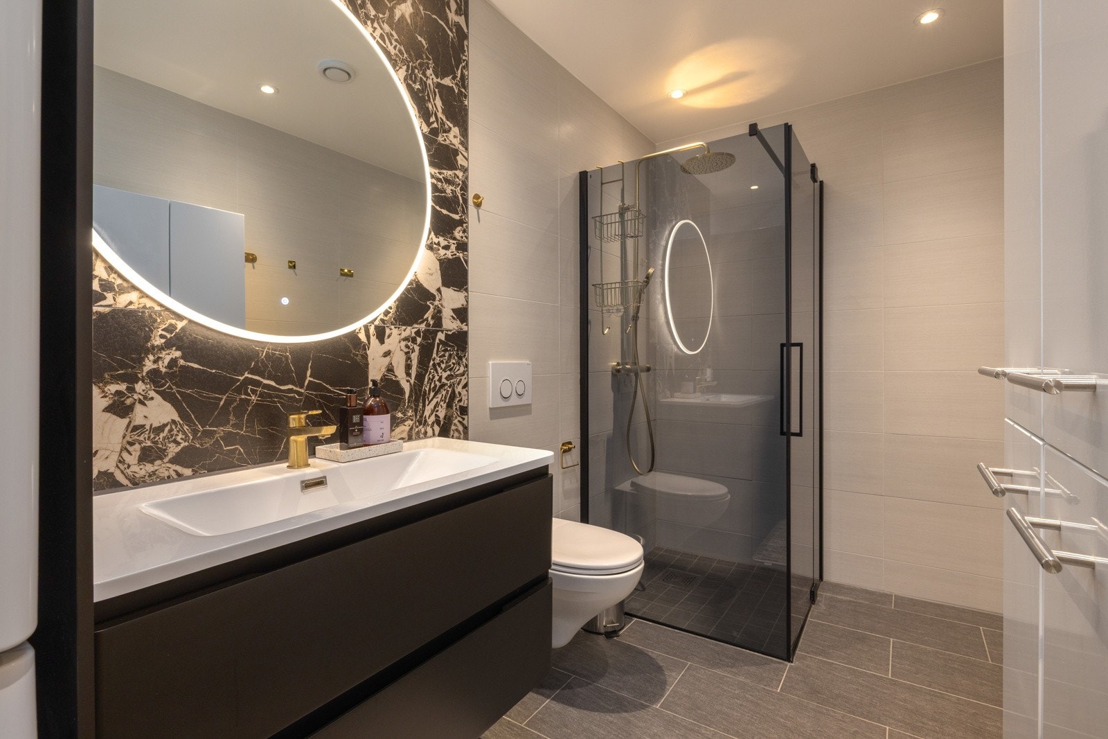 Baderom med rund speilbelysning, mørke kontraster og dusjhjørne i glass. Galleribilde