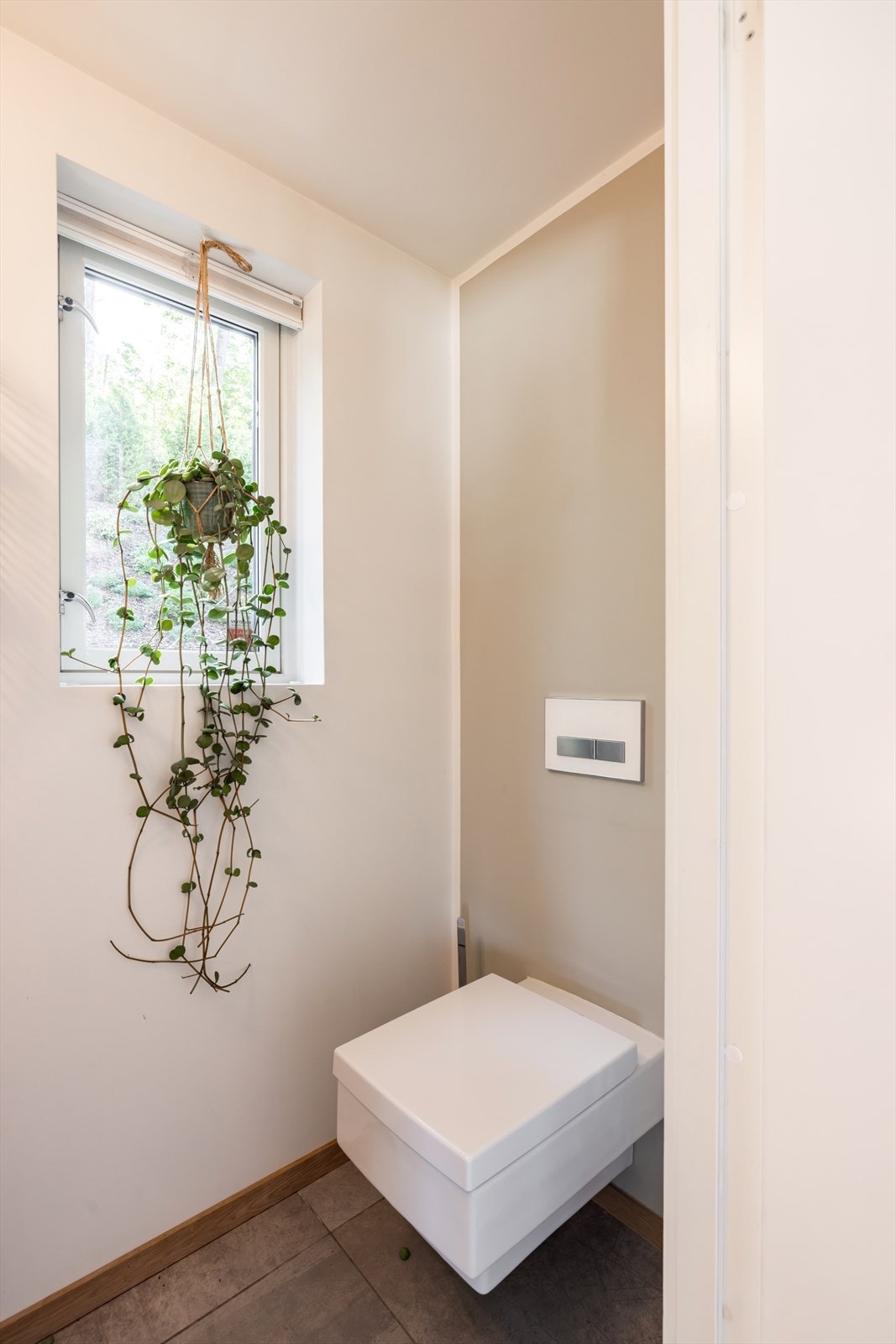 Praktisk toalettrom i 1. etasje med veggmontert toalett, servant, speil og vindu mot terrassen på baksiden av boligen. Galleribilde