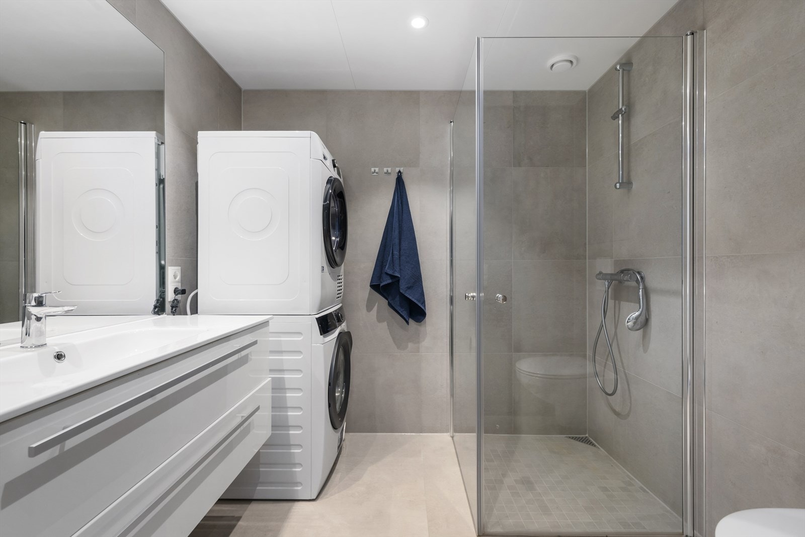 Moderne og funksjonelt baderom med grå fliser, dusjhjørne med glassvegg, servant med underskap og speil, samt praktisk vaskesøyle med vaskemaskin og tørketrommel. Galleribilde