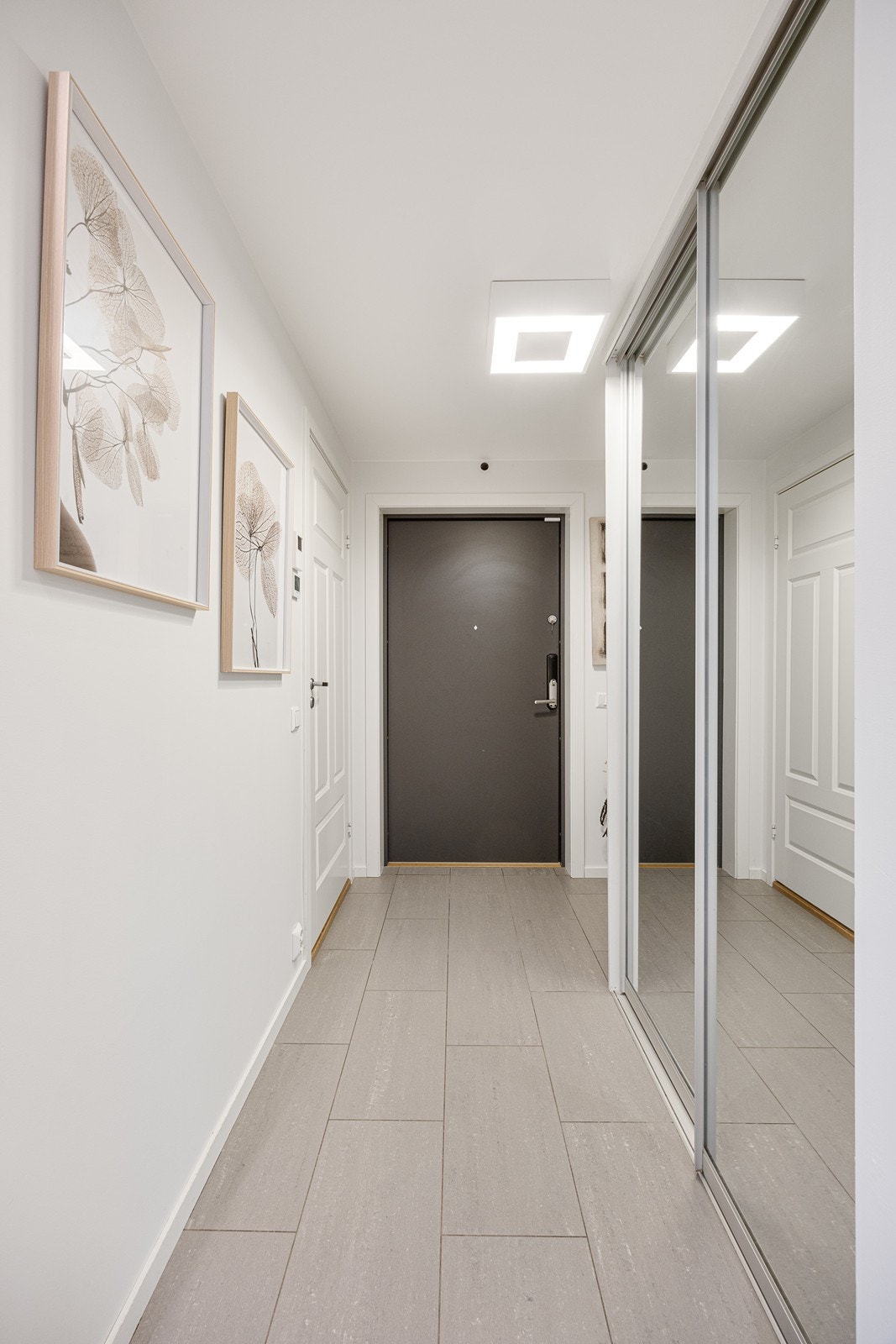 Entré med hvitmalte vegger og lysegrå fliser på gulv med vannbåren gulvvarme. Yttertøyet ryddes enkelt bort i en plassbygd skyvedørsgarderobe med speilfronter Galleribilde