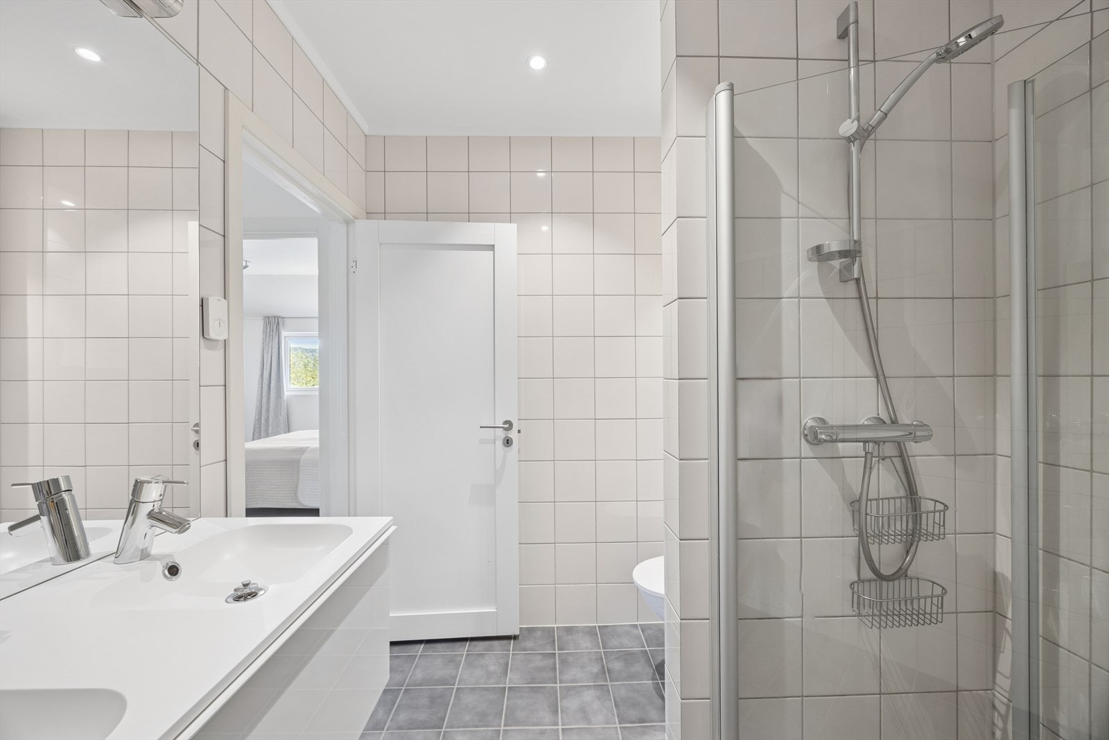 Badet er utstyrt med vegghengt toalett, dusjhjørne med svingbare glassdører, samt baderomsinnredning med to nedfelte servanter. Galleribilde