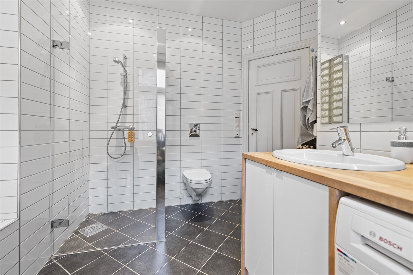 Dusjnisje m/glassdør, spesialbygd innredning i hvitoljet eik benkeplate (Kvik) og servant med speil. Veggmontert wc. Galleribilde