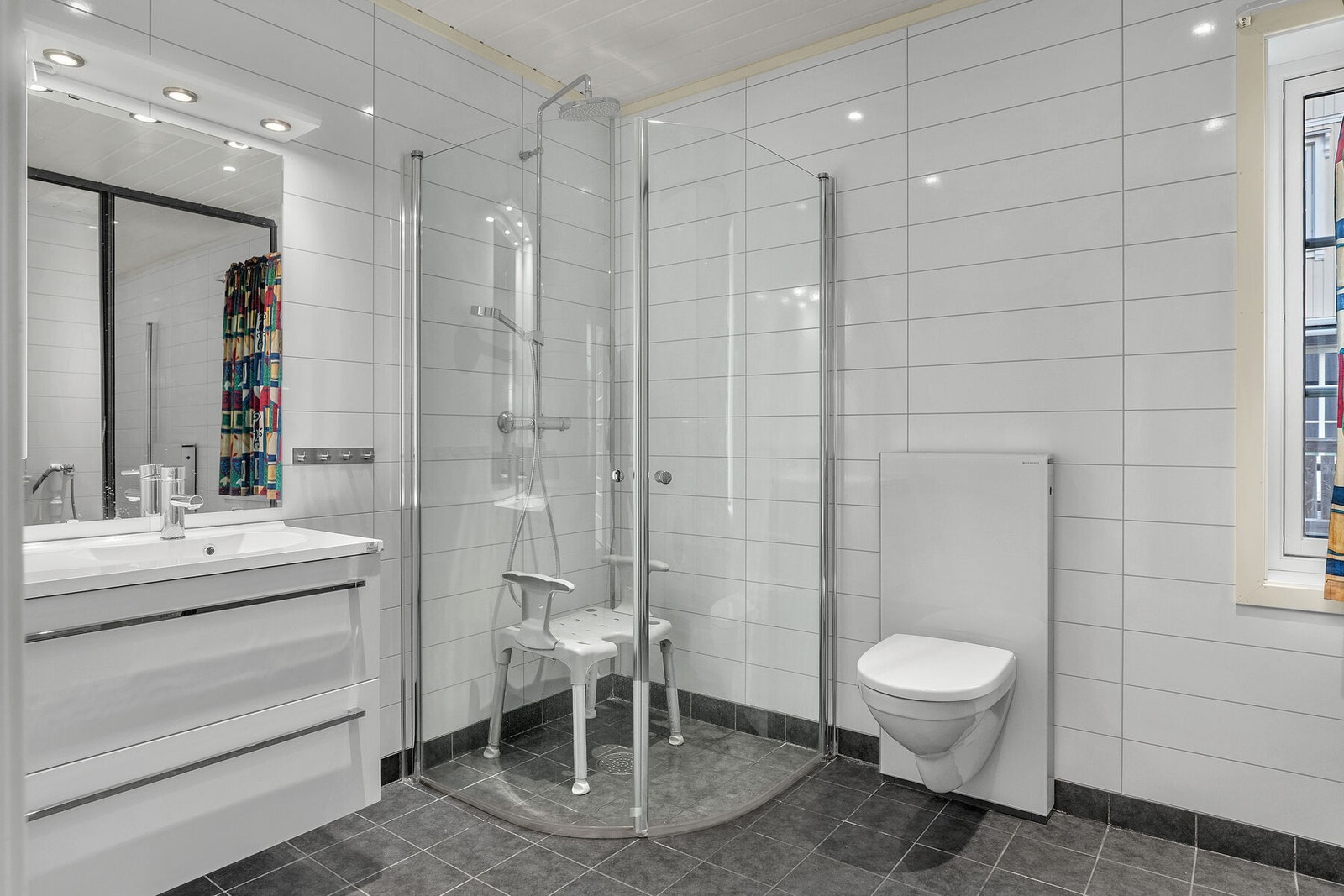 Bad/vaskerom med servant på innredning, veggmontert toalett og dusjvegger. Rommet har flislagt gulv med elektrisk gulvvarme. Galleribilde