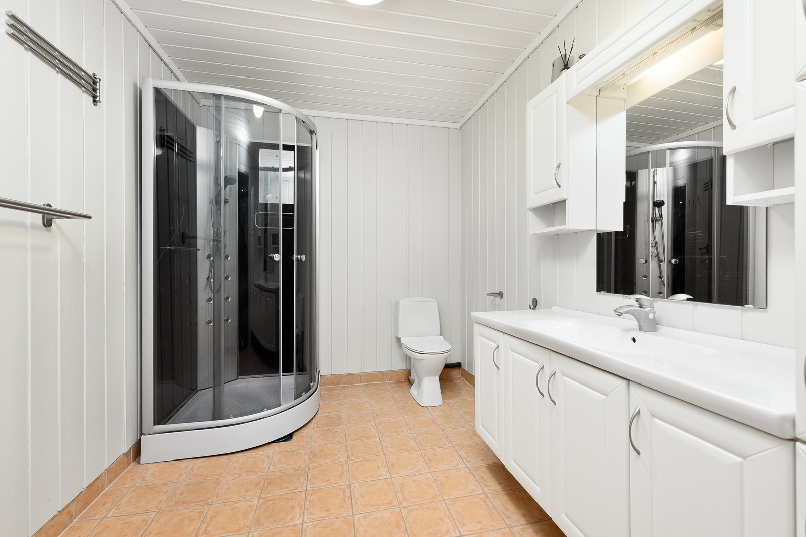 Badet har malt panel på vegger og flislagt gulv. I badet er det montert servantinnredning med skap, lukket dusjkabinett og frittstående toalett. Galleribilde